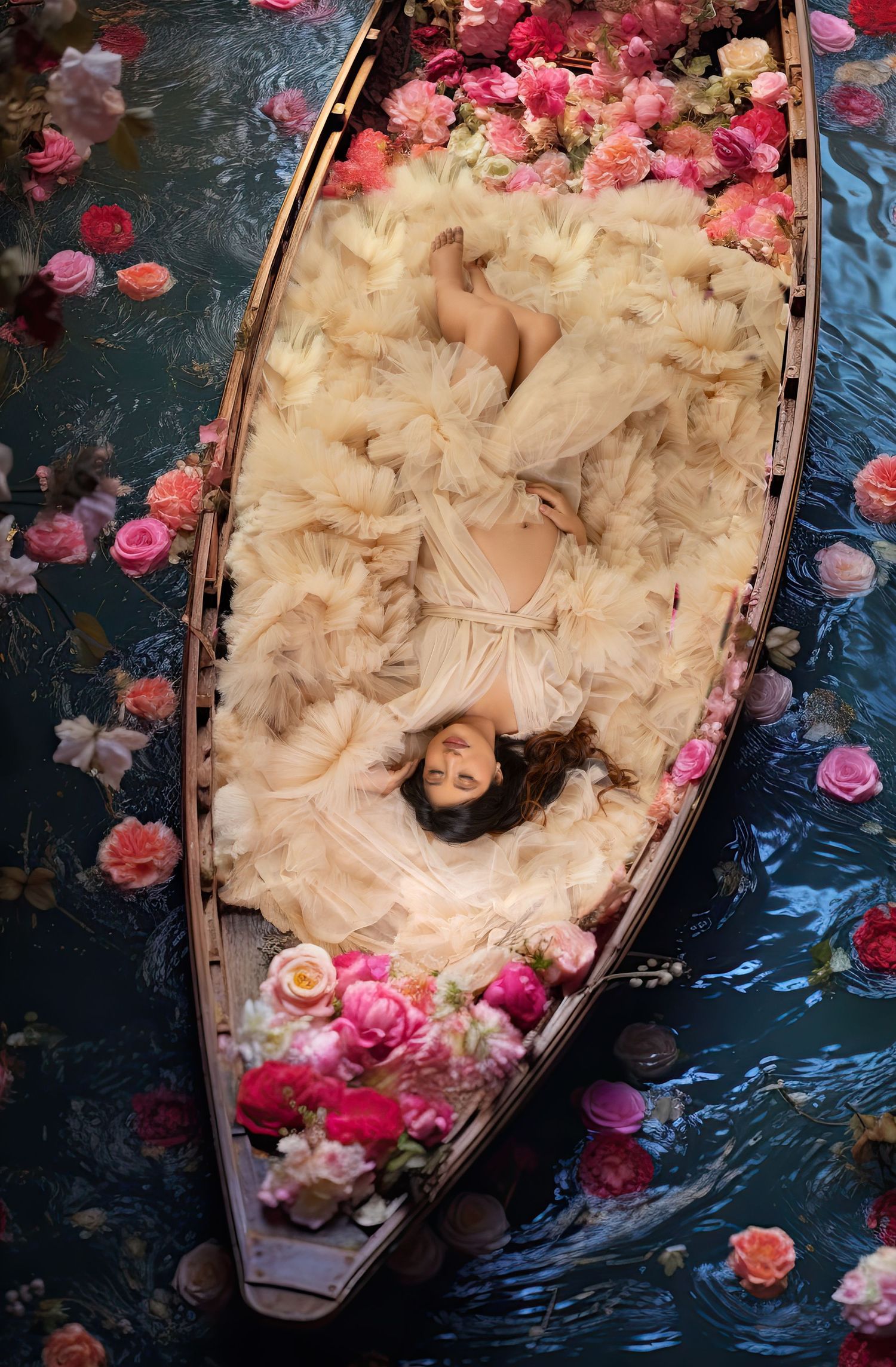 Aerial view of pregnant woman in white tulle gown lying in flower-filled boat on calm water.