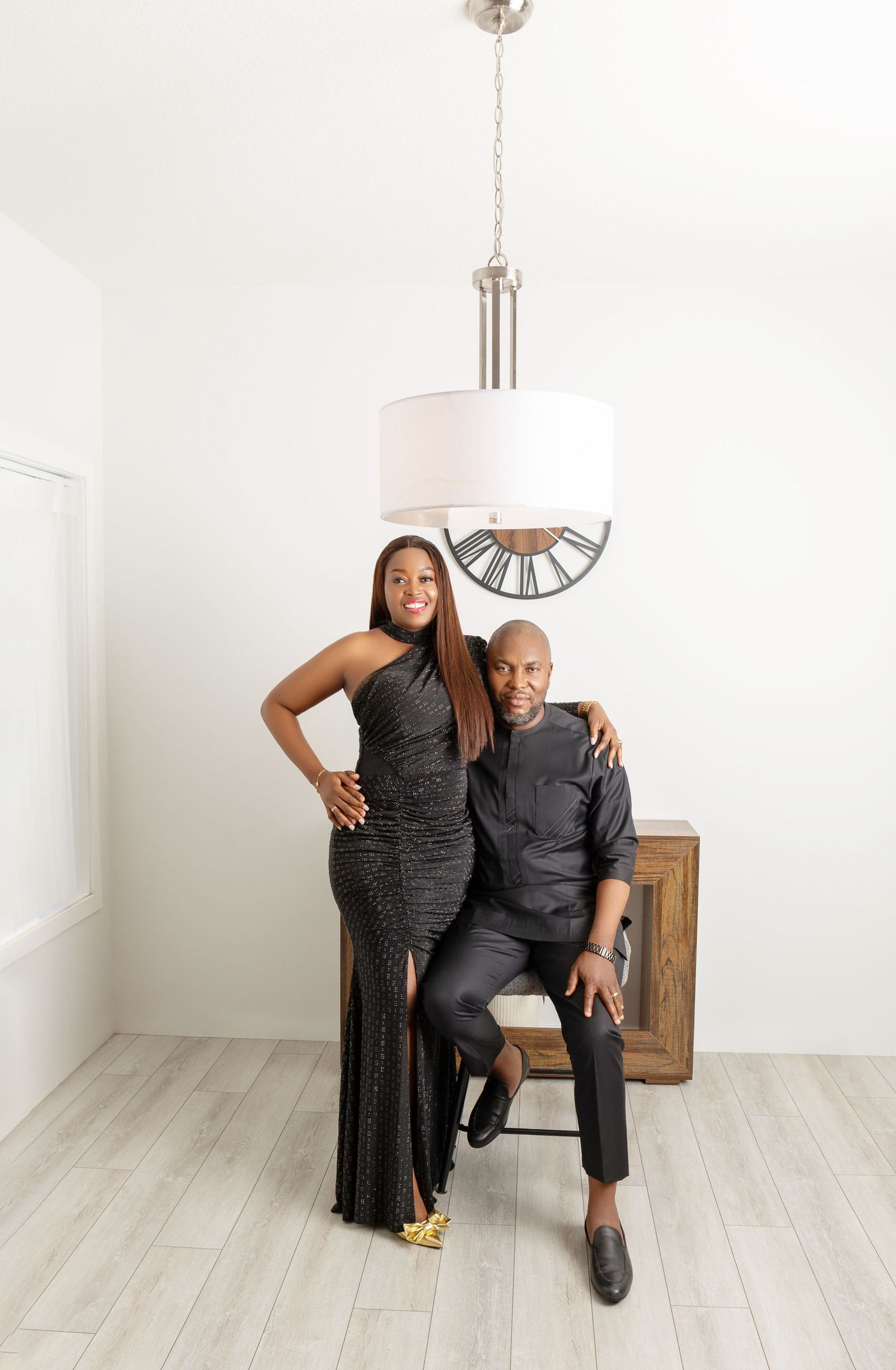 Elegant couple in matching black formal attire posing under a pendant light in a bright modern interior.