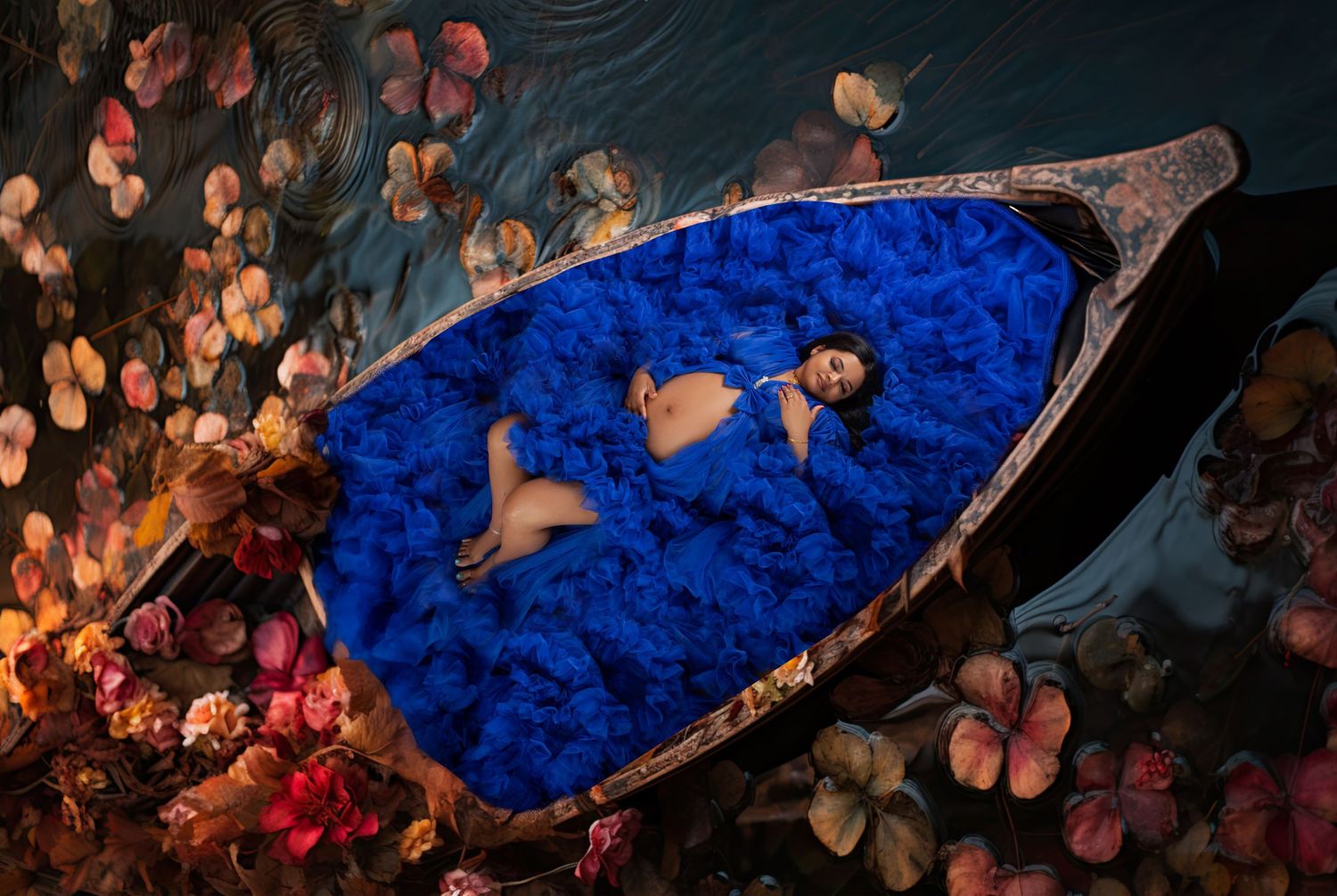 Woman in bikini reclining on blue feathers in a wooden boat surrounded by floating flowers on dark water.