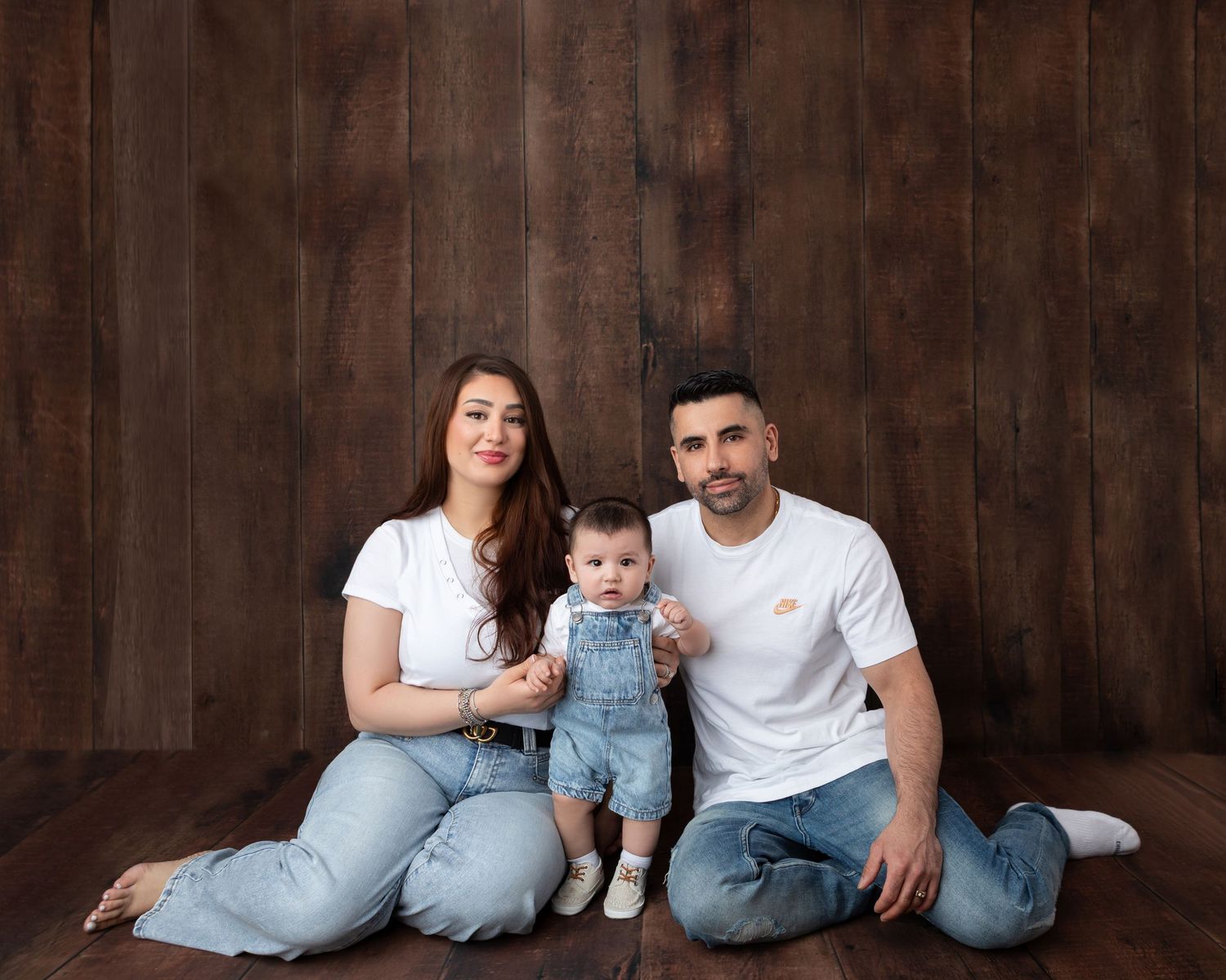 A young family with a baby in denim overalls poses on the floor against a rustic wood backdrop.
