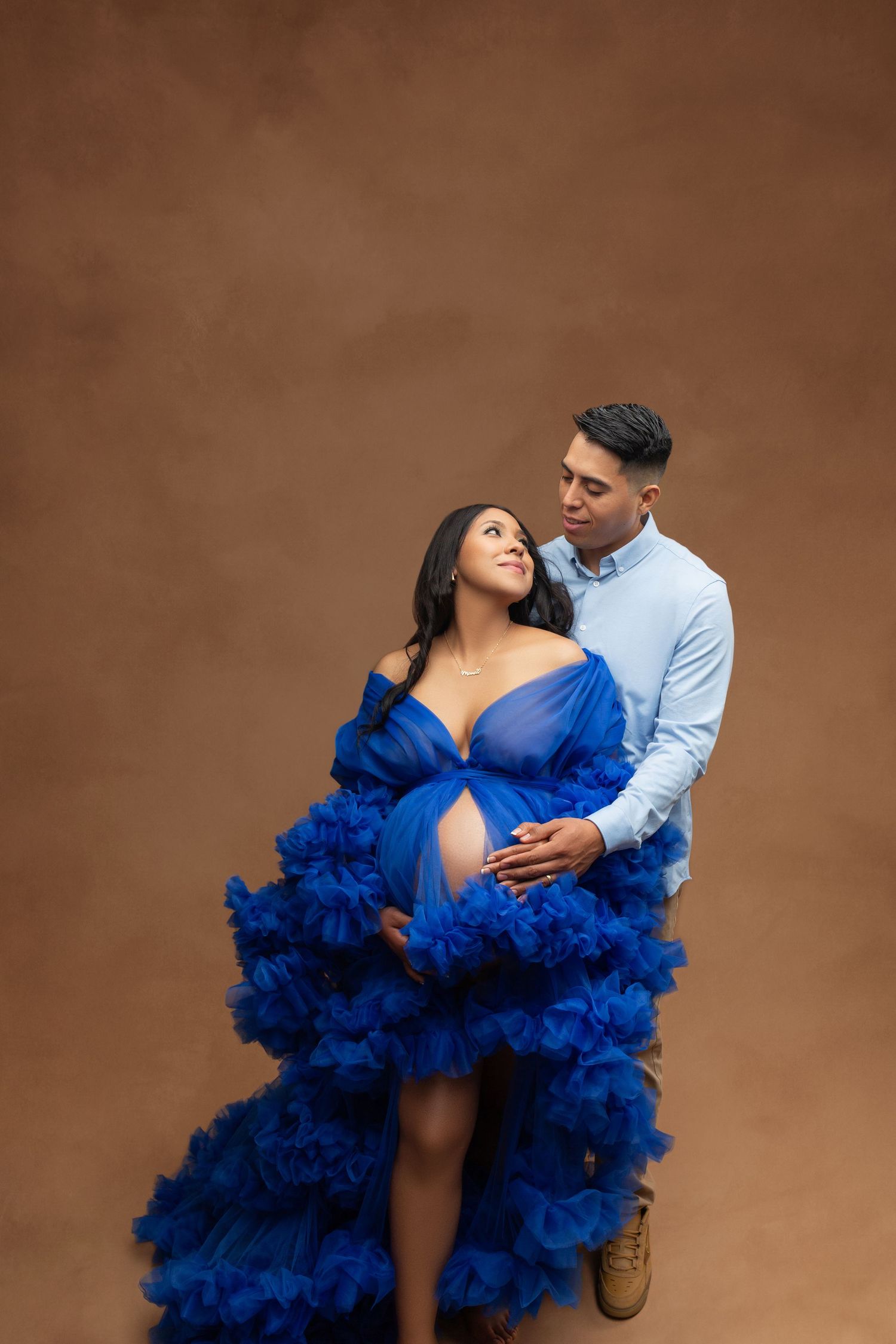 Couple in maternity photo shoot, woman in royal blue tulle gown cradling baby bump, man in light blue shirt embracing her.