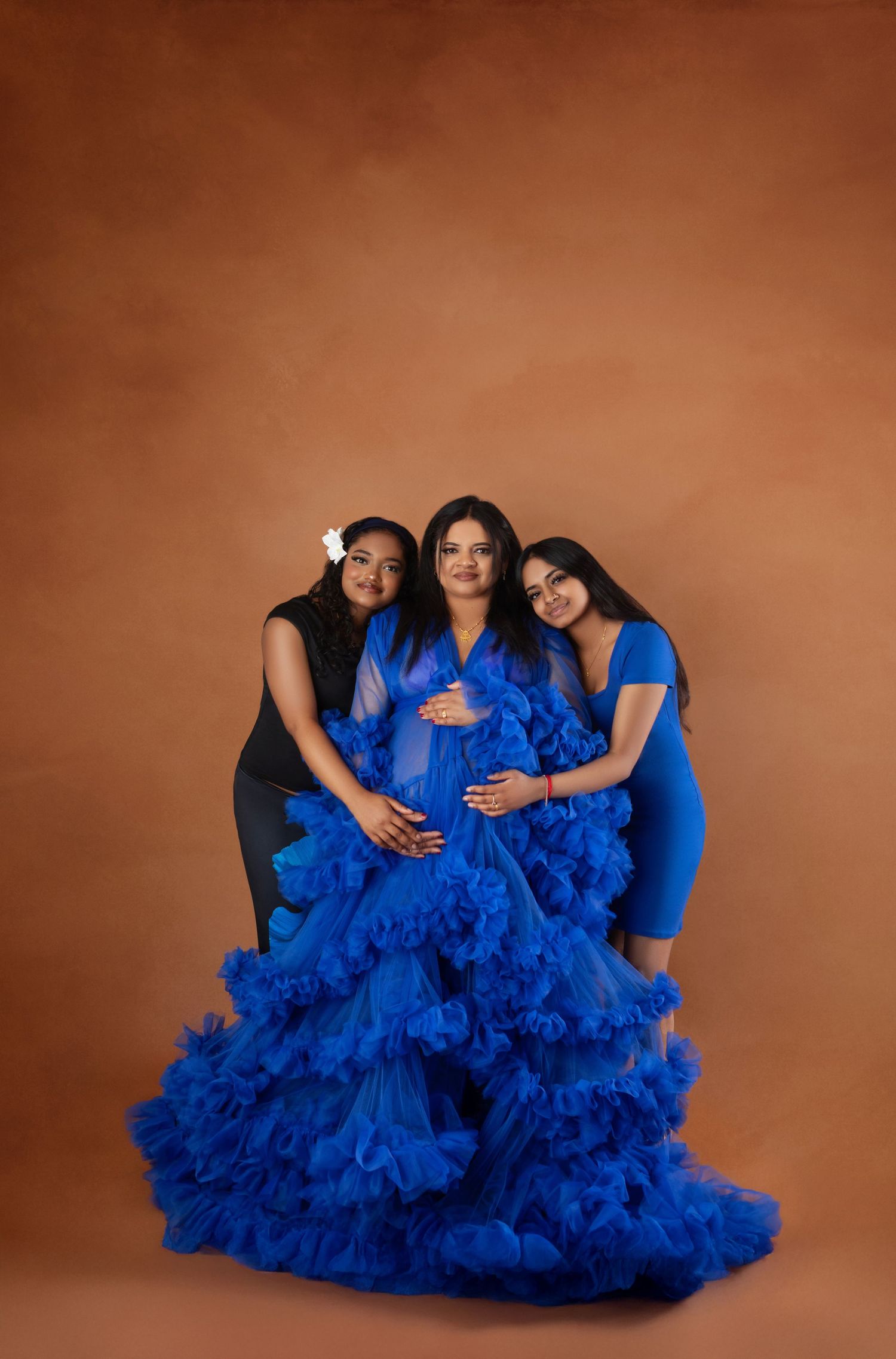 Three women pose together in a studio, the center wearing a stunning royal blue ruffled maternity gown.