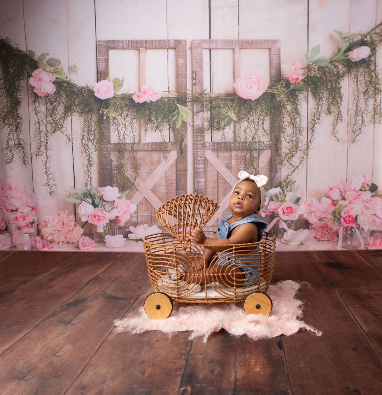 Baby girl in blue dress sits in wicker cart on wood floor with floral barn door backdrop.