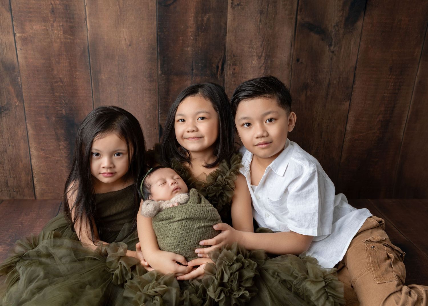 Three siblings pose with a newborn baby wrapped in green, sitting against a rustic wood backdrop in a studio portrait.