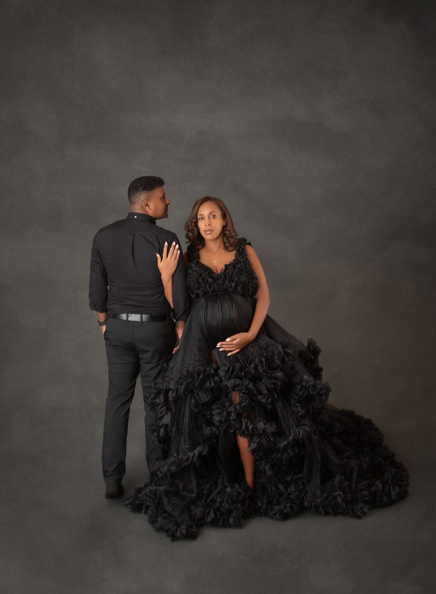 Couple posing in studio maternity shoot, pregnant woman in dramatic black ruffled gown with partner in black attire.