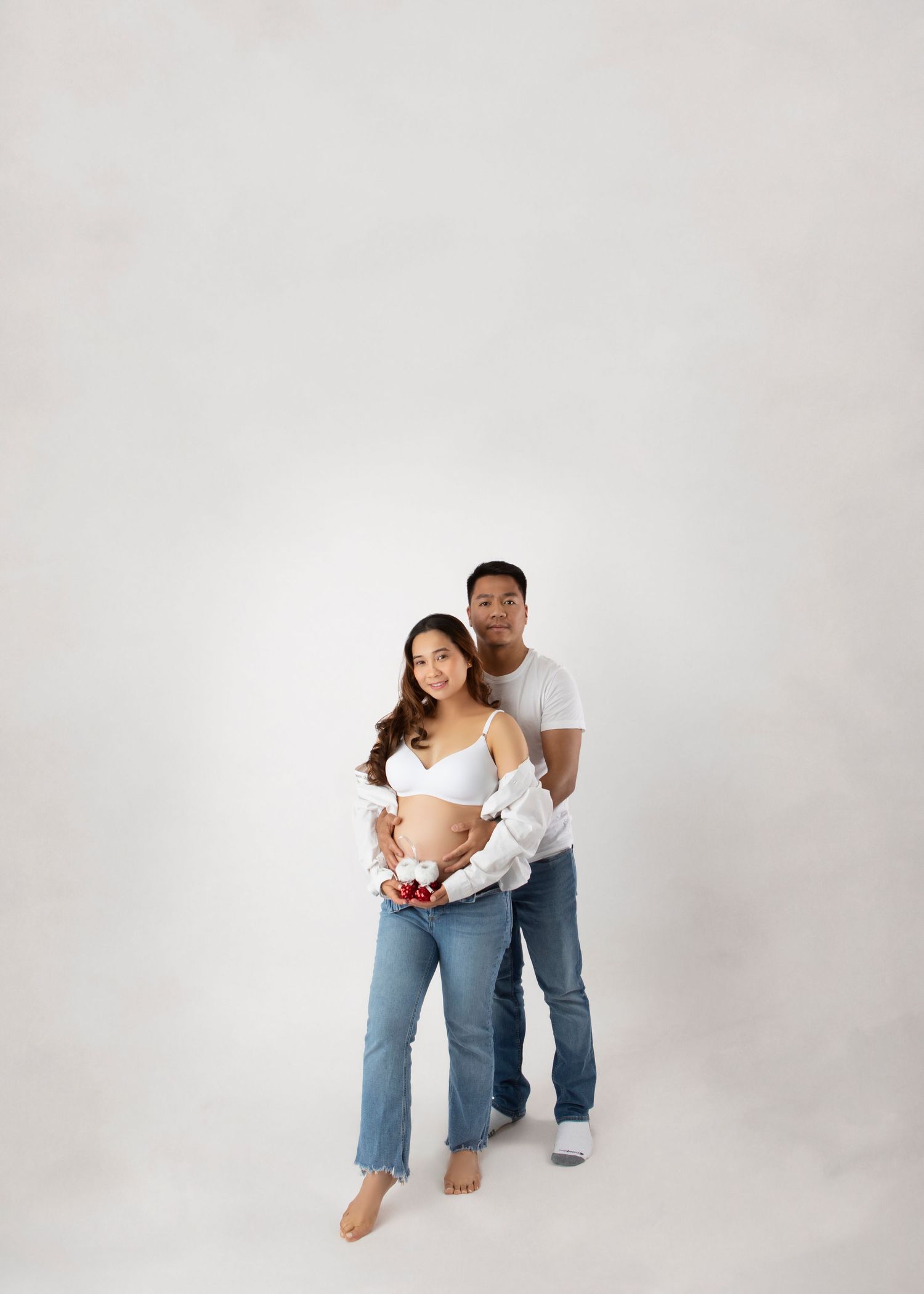Full length portrait of smiling pregnant couple in white and denim holding tiny red baby shoes.
