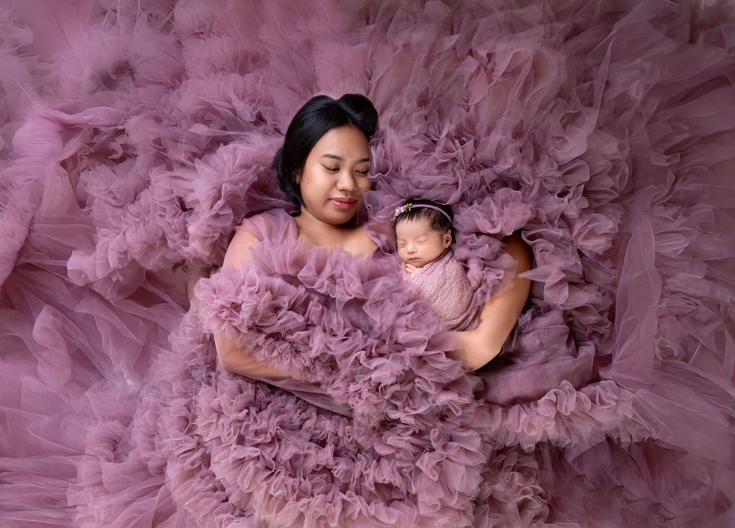 Mom in purple ruffle gown lying with sleeping swaddled newborn baby in professional photo shoot.