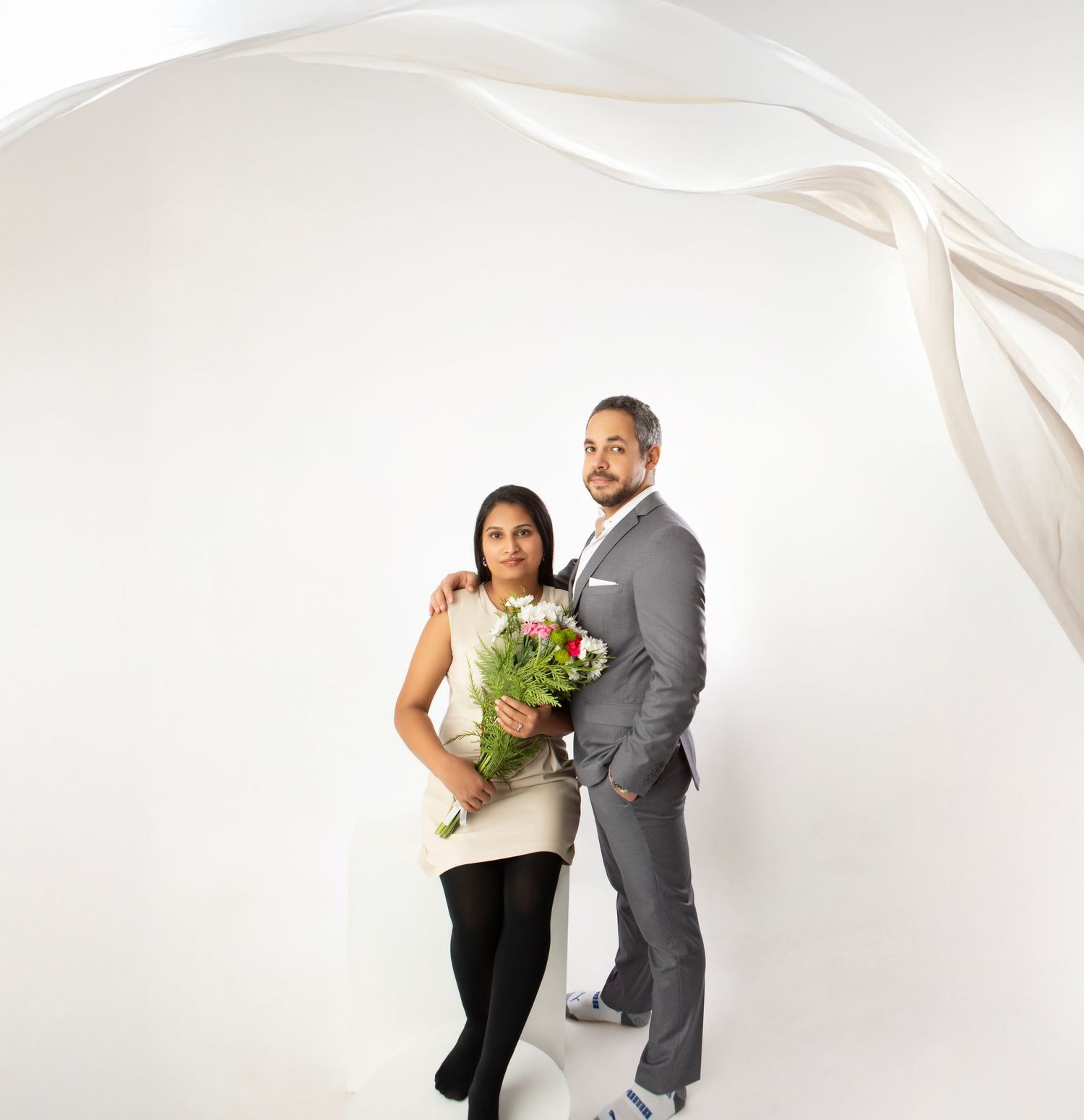 Couple standing together holding red and white flowers with flowing fabric backdrop in white studio.