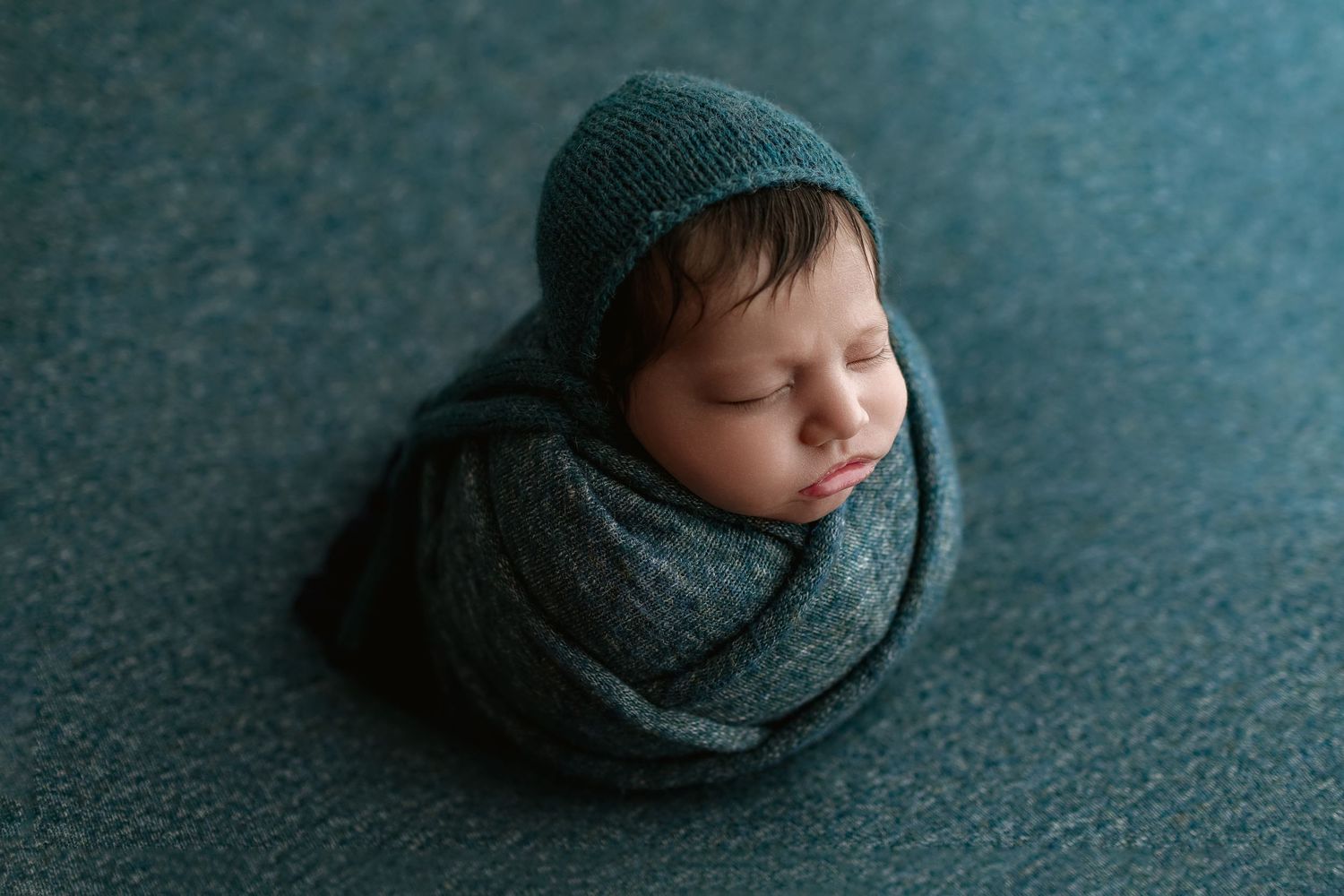 Newborn baby wrapped in teal knit blanket with matching bonnet, sleeping peacefully in a cozy swaddle pose.