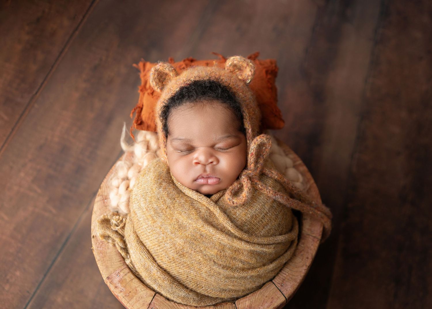 Sleeping newborn baby wrapped in a tan swaddle wearing a bear bonnet in a wooden bowl.