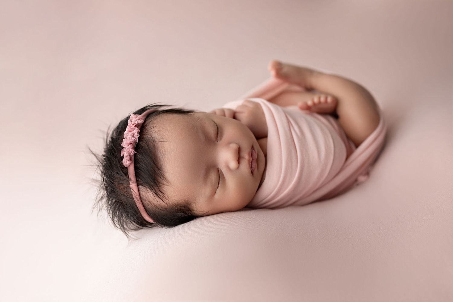 Sleeping newborn baby girl swaddled in pink wrap with floral headband on soft pink background.
