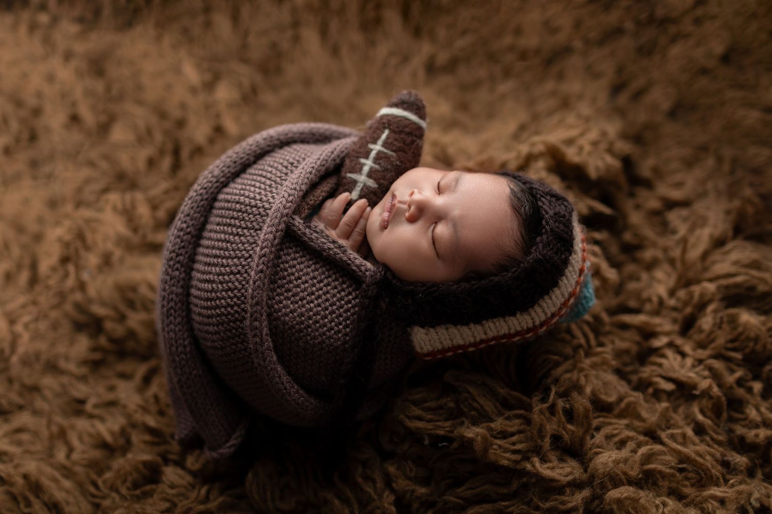 Sleeping newborn baby wrapped in brown knit outfit holding a tiny football on a fluffy brown fur rug.