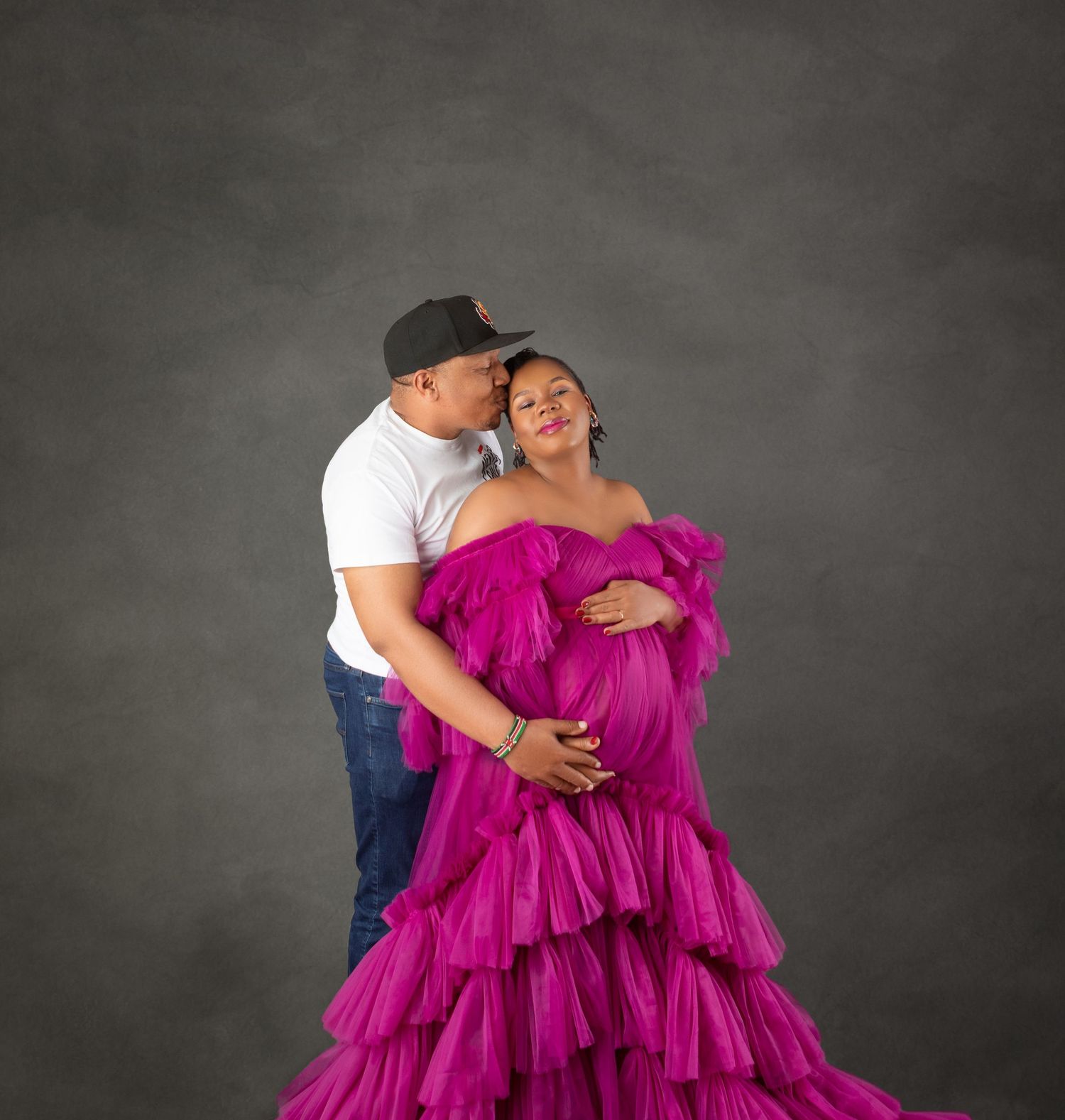 Pregnant woman in magenta ruffle gown poses with partner kissing her cheek during maternity photo session.