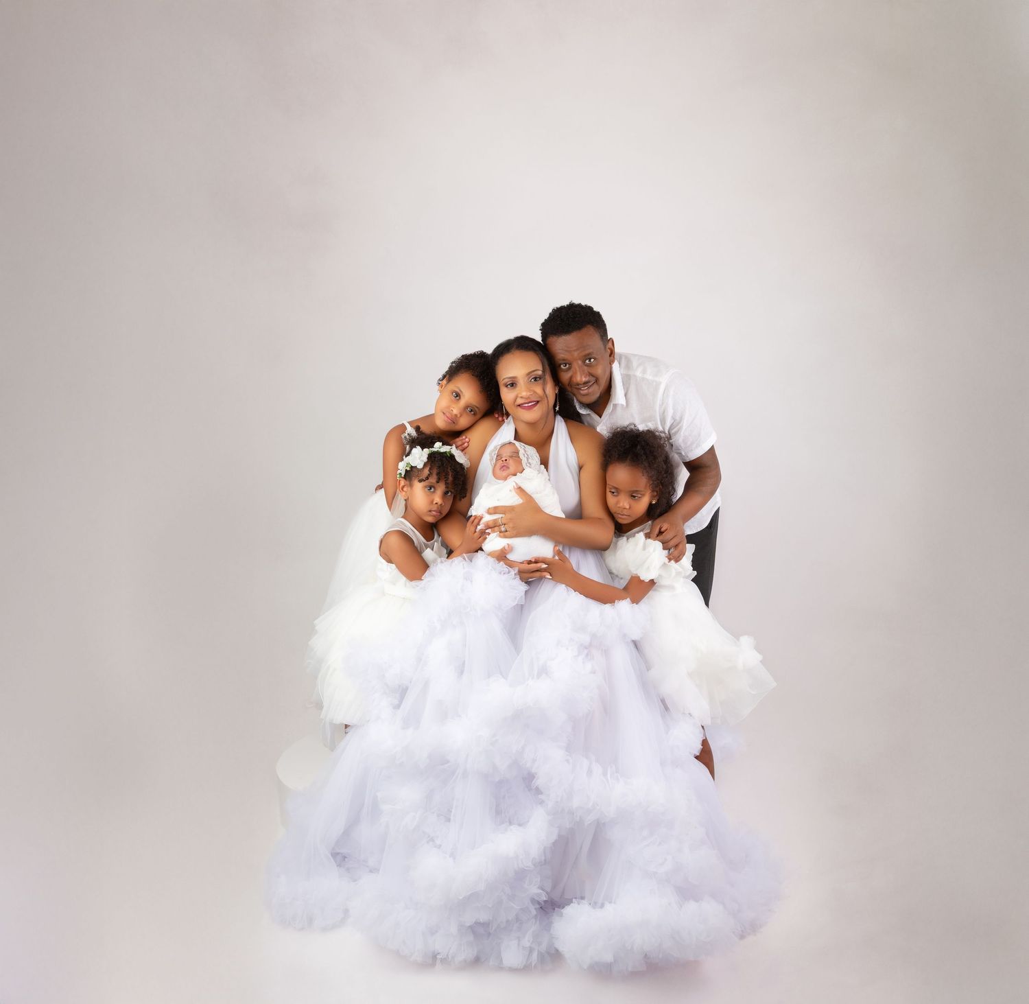 A joyful family in white outfits poses together, with children hugging parents against a soft white background.