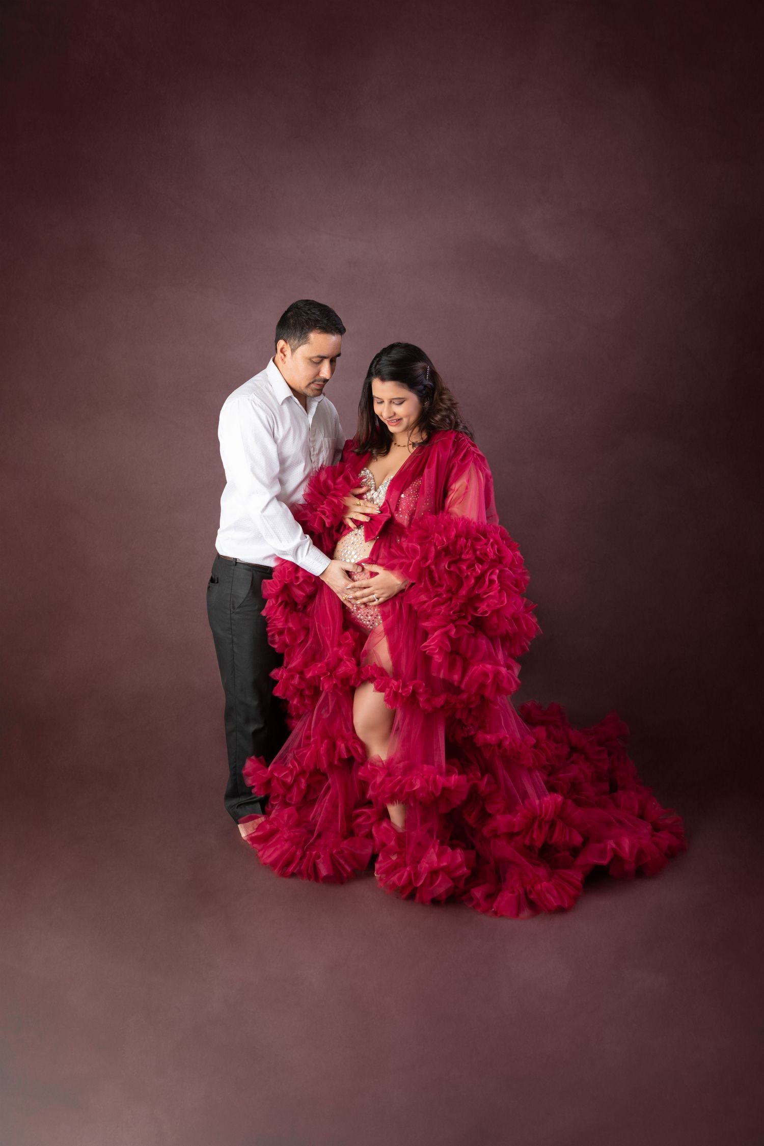 Couple maternity photo with pregnant woman in red ruffled gown and man in white shirt on brown backdrop.