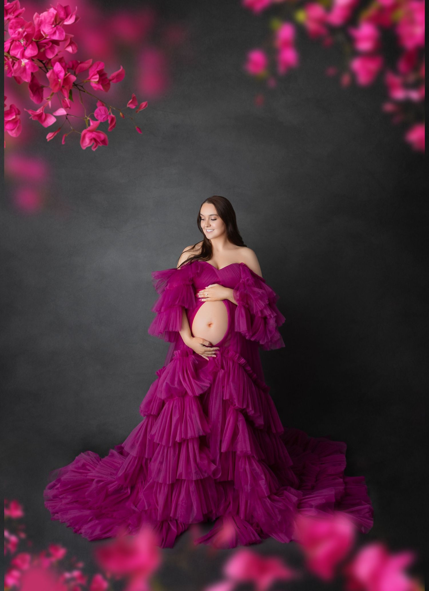 Pregnant woman in vibrant magenta ruffled tulle gown surrounded by pink flowers in studio setting.