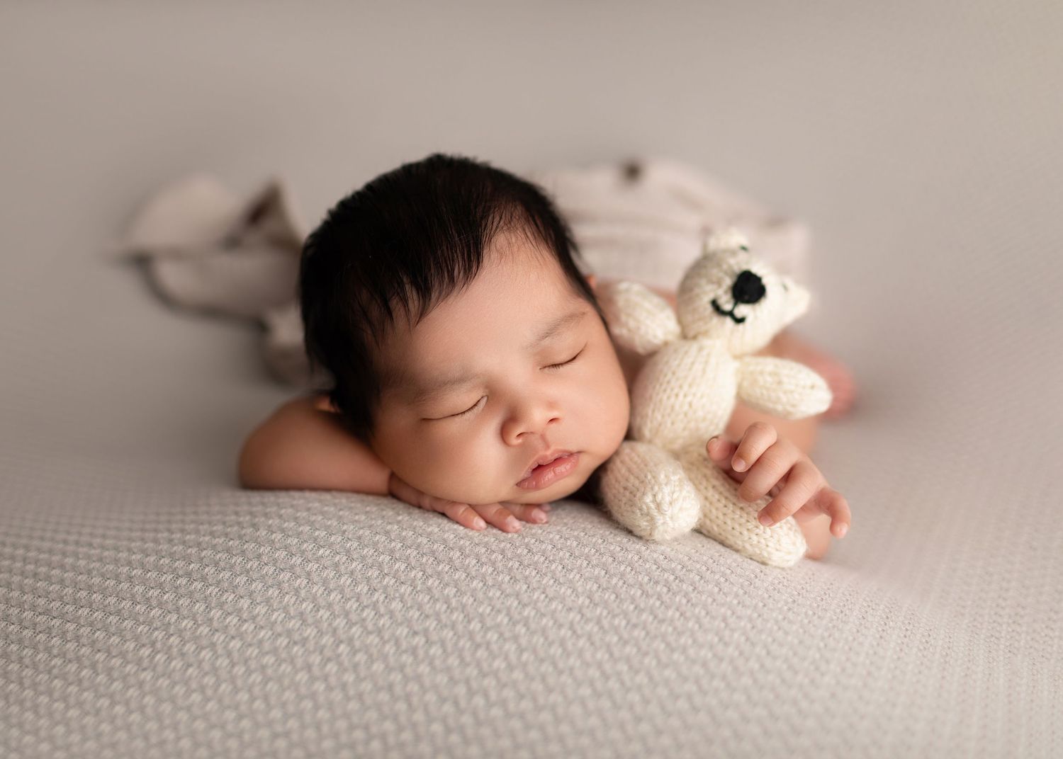Sleeping newborn baby holding a knitted white teddy bear on a soft gray textured blanket.