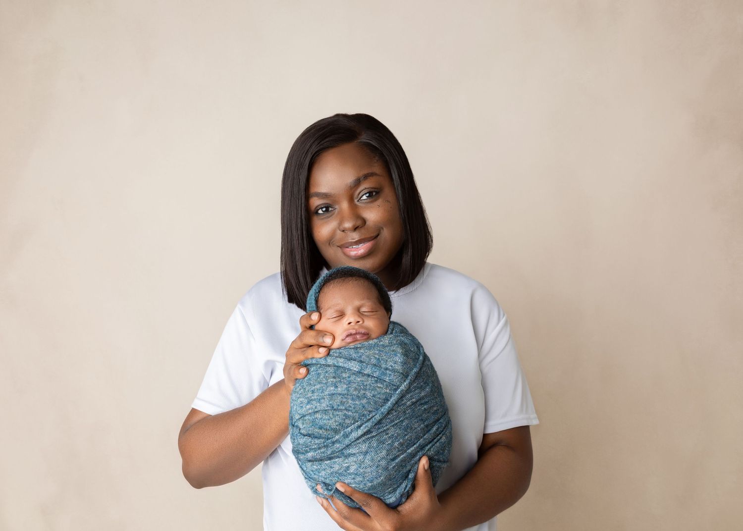 Young mother in white shirt proudly holding teal-swaddled newborn baby against warm beige studio backdrop.