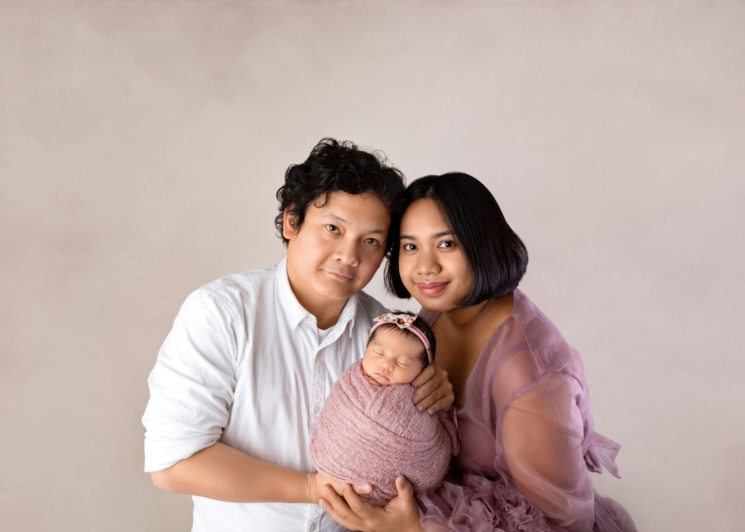 Asian family portrait with parents in white and purple holding pink-swaddled newborn against neutral backdrop.