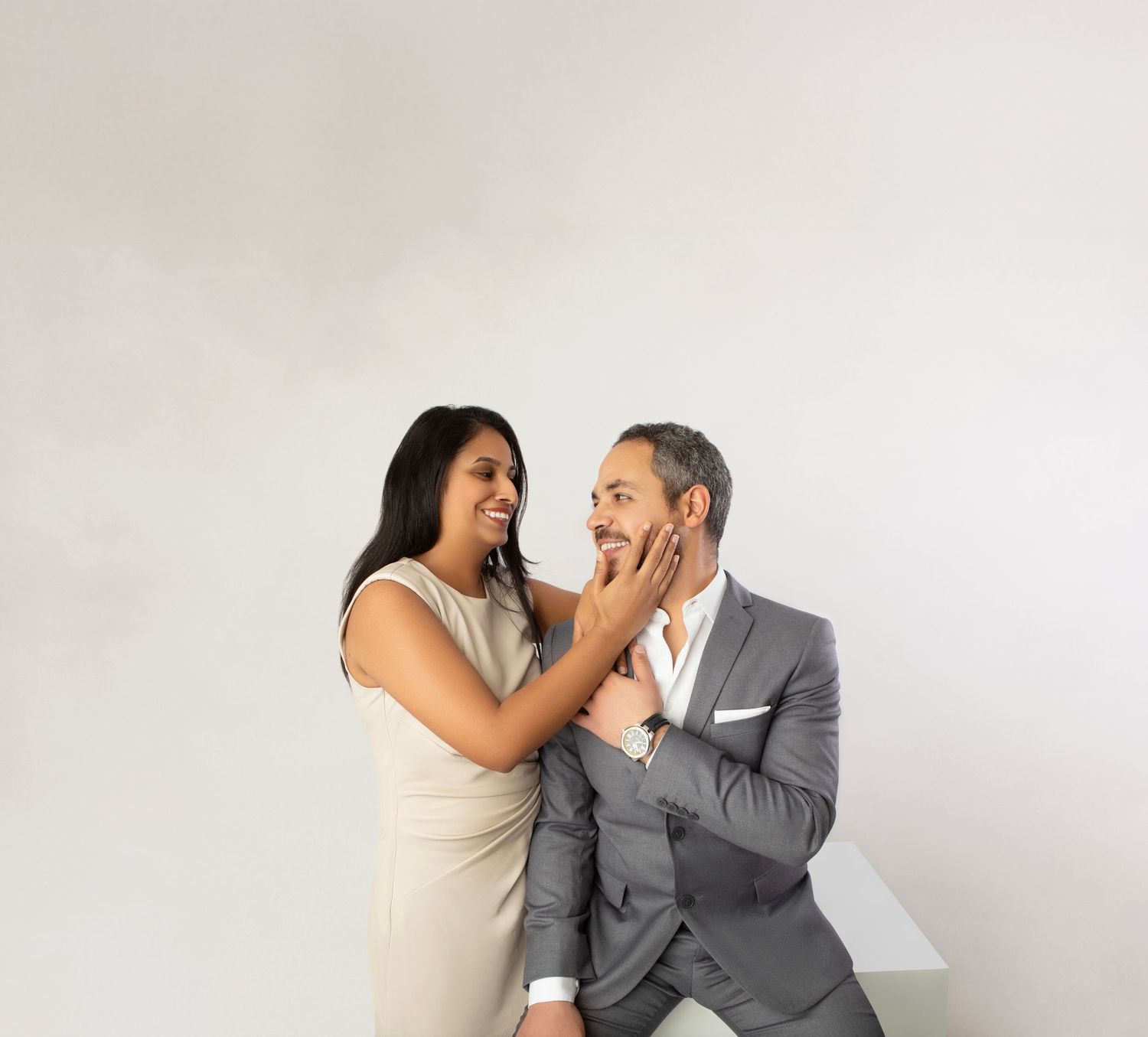 Loving interracial couple smiling at each other, woman touching mans face, man in gray suit, white background.