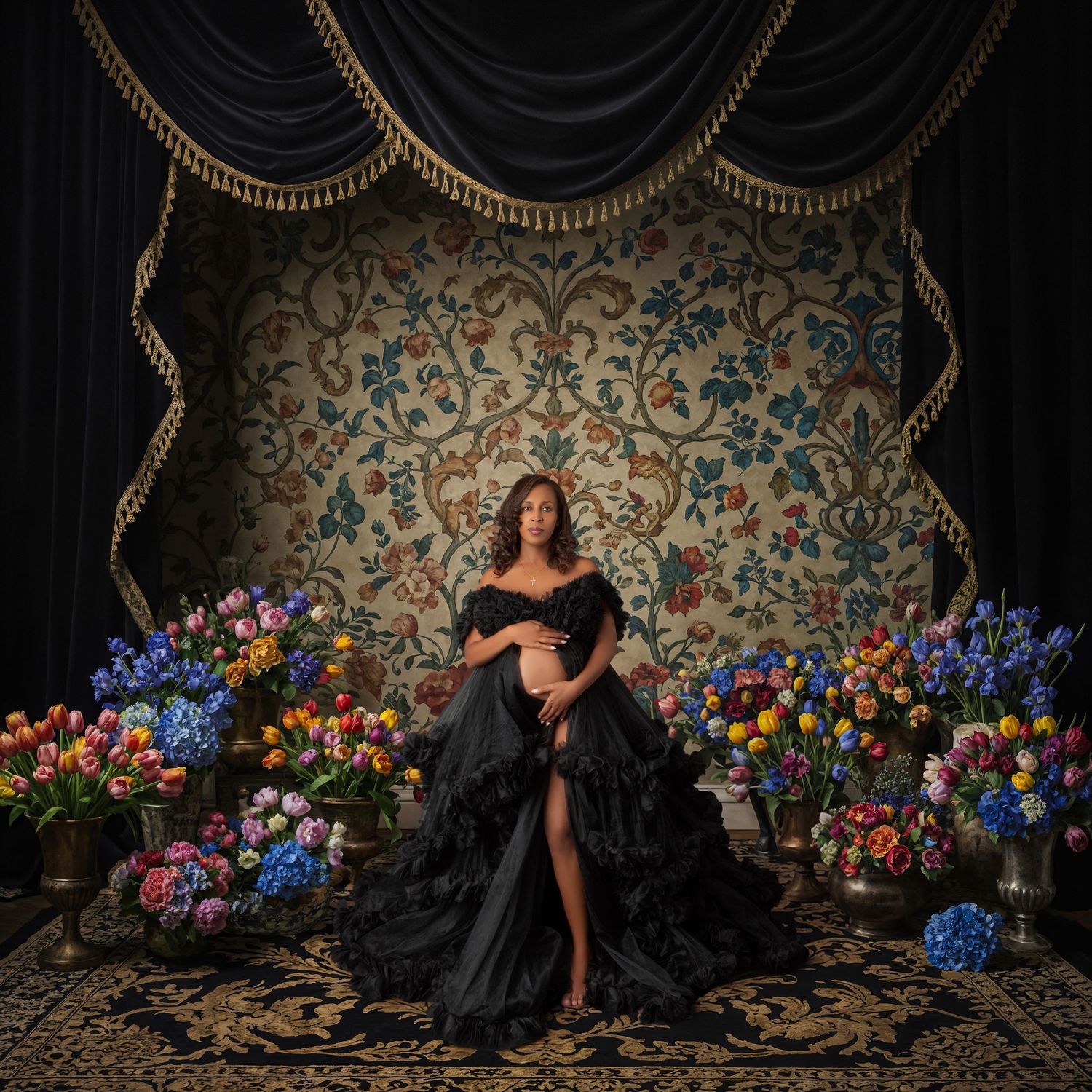 Pregnant woman in black ruffled gown poses regally among colorful floral arrangements against ornate tapestry backdrop.