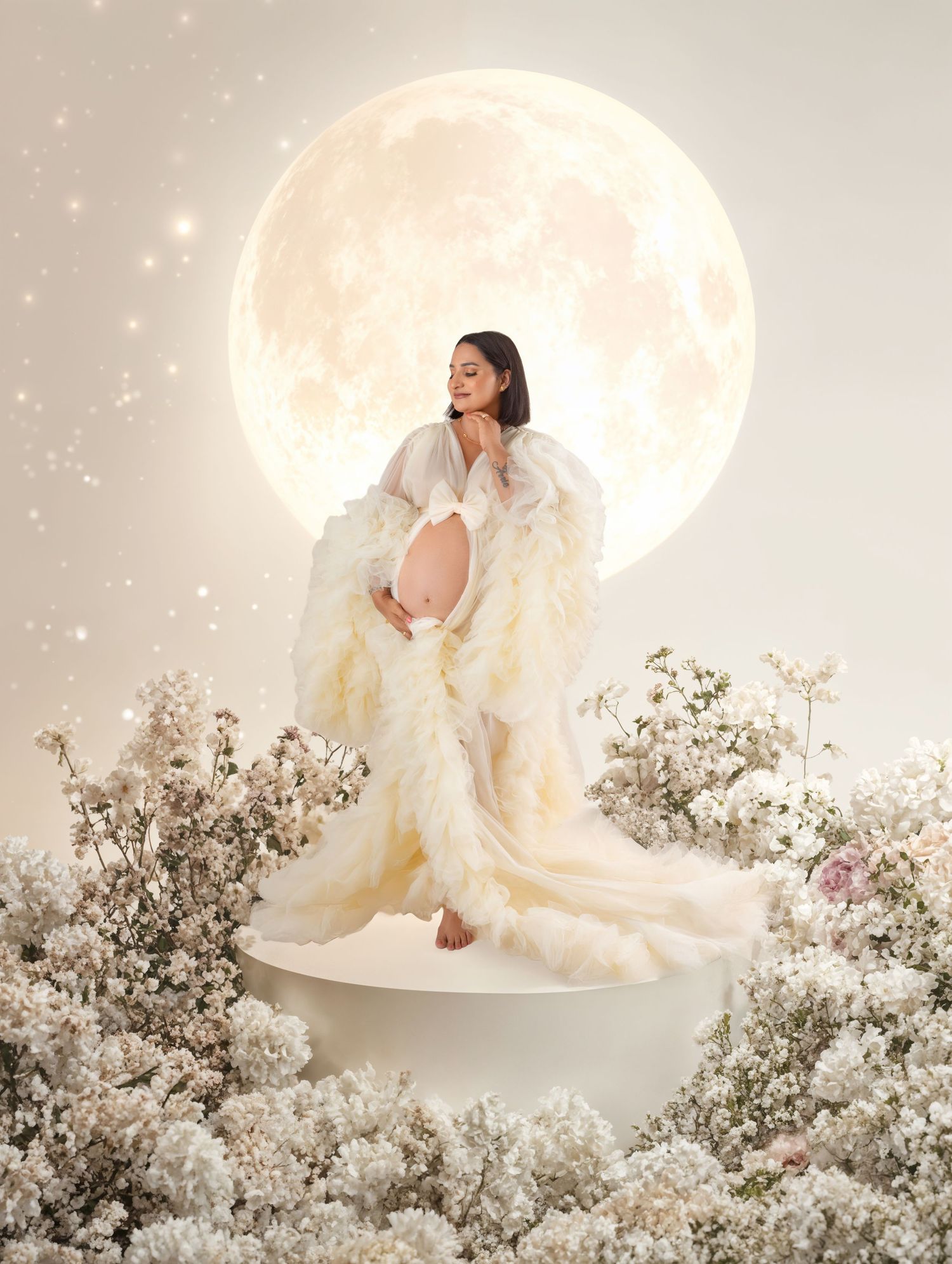 Pregnant woman in white ruffled gown posed before glowing full moon surrounded by delicate white flowers.
