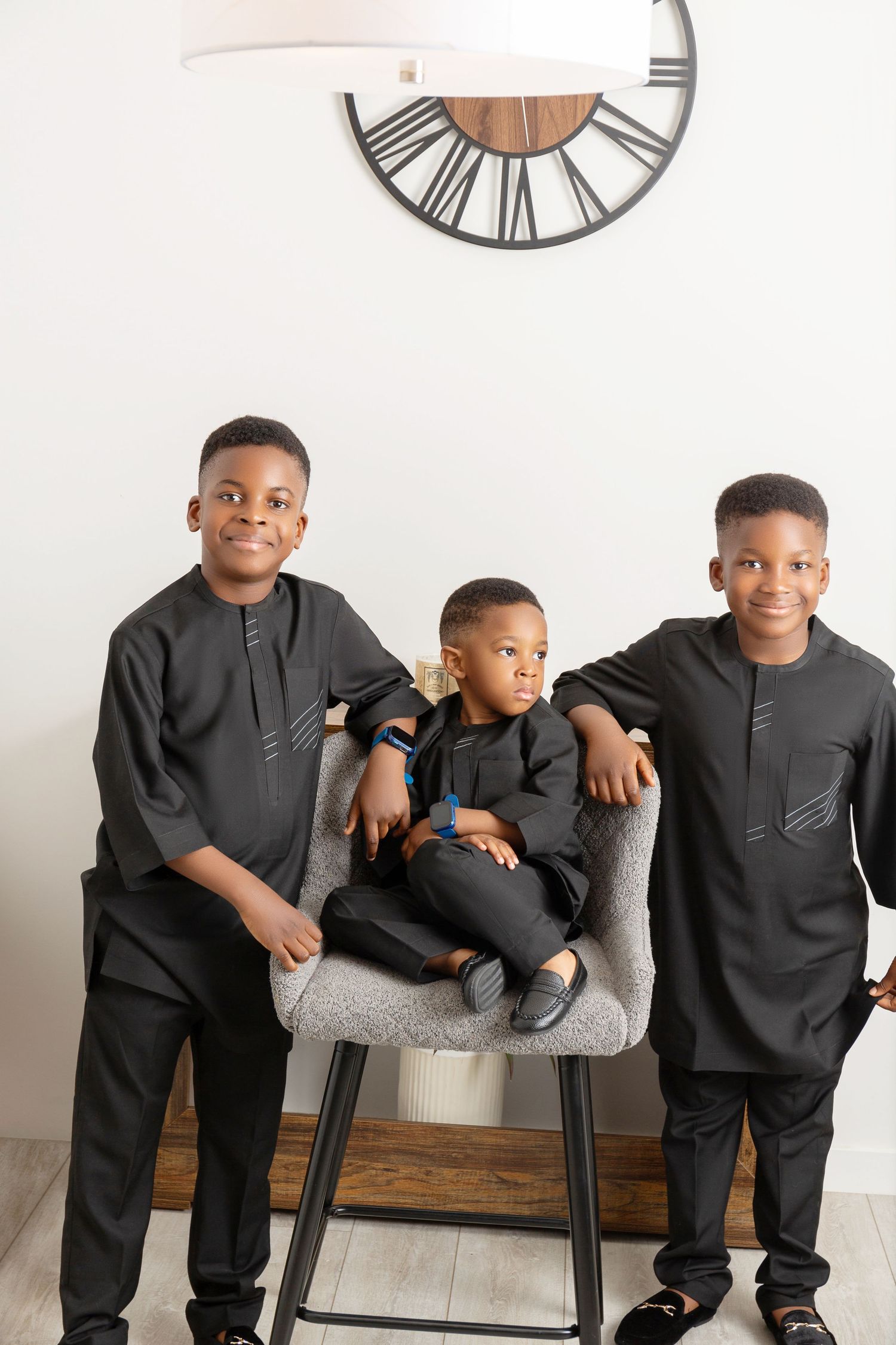 Three young boys in matching black traditional African outfits pose together near a decorative clock.
