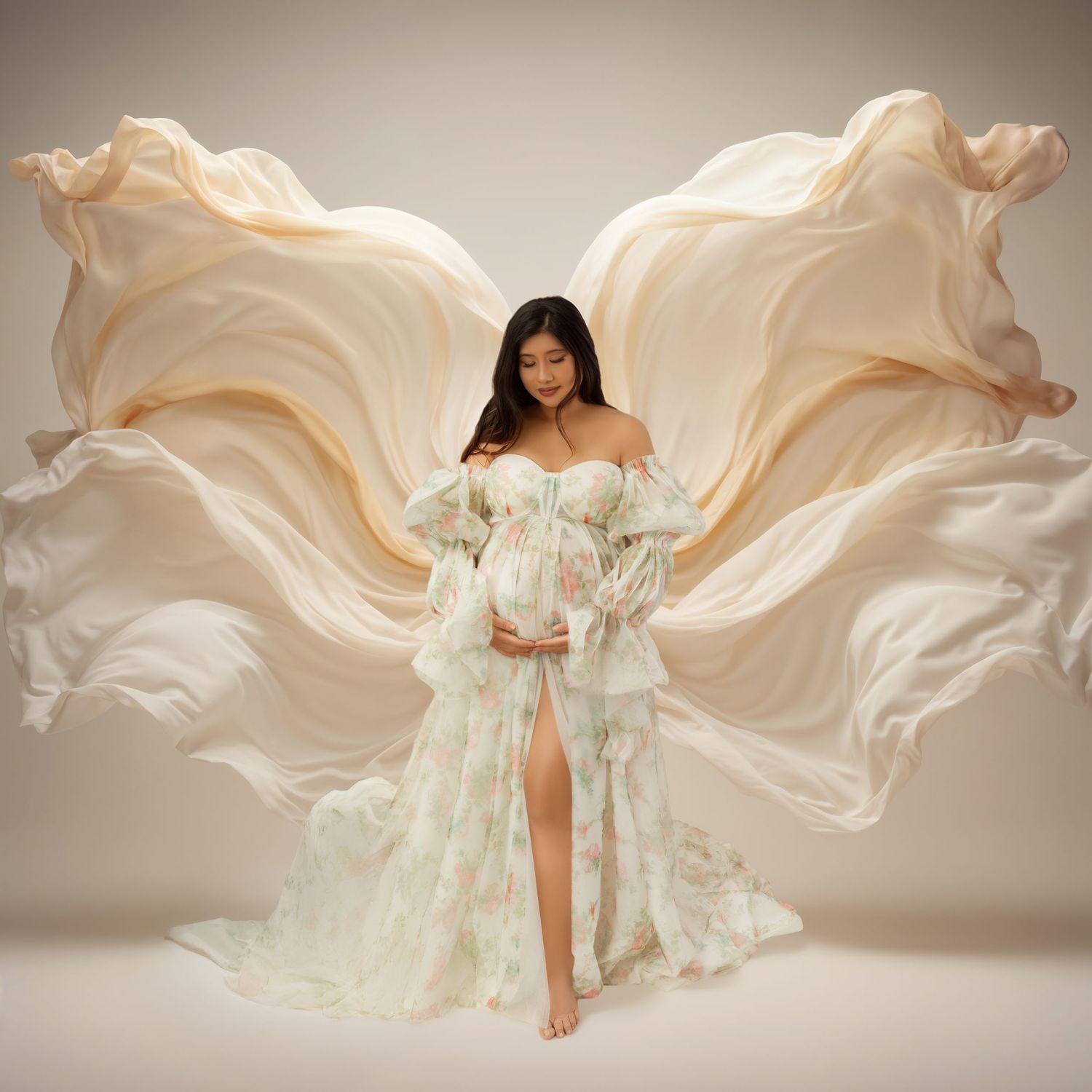 Pregnant woman in floral gown with flowing cream fabric wings in elegant maternity photo shoot.