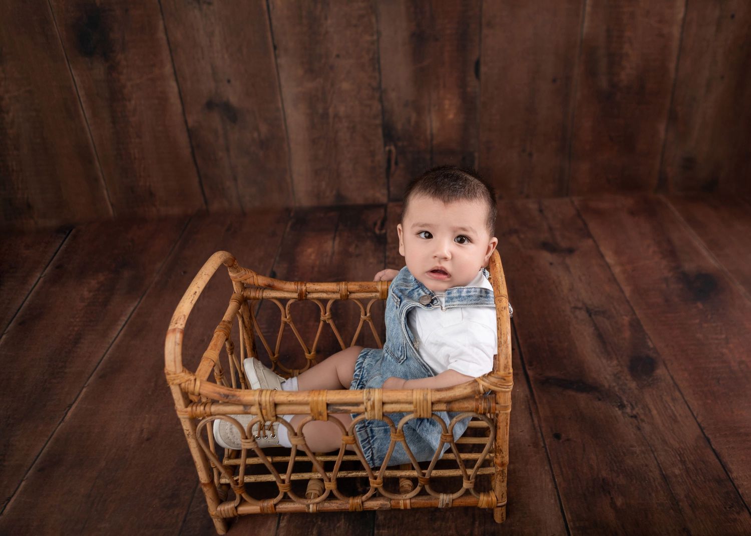 A cute baby in denim overalls sitting in a wicker cradle on a rustic wooden floor backdrop.