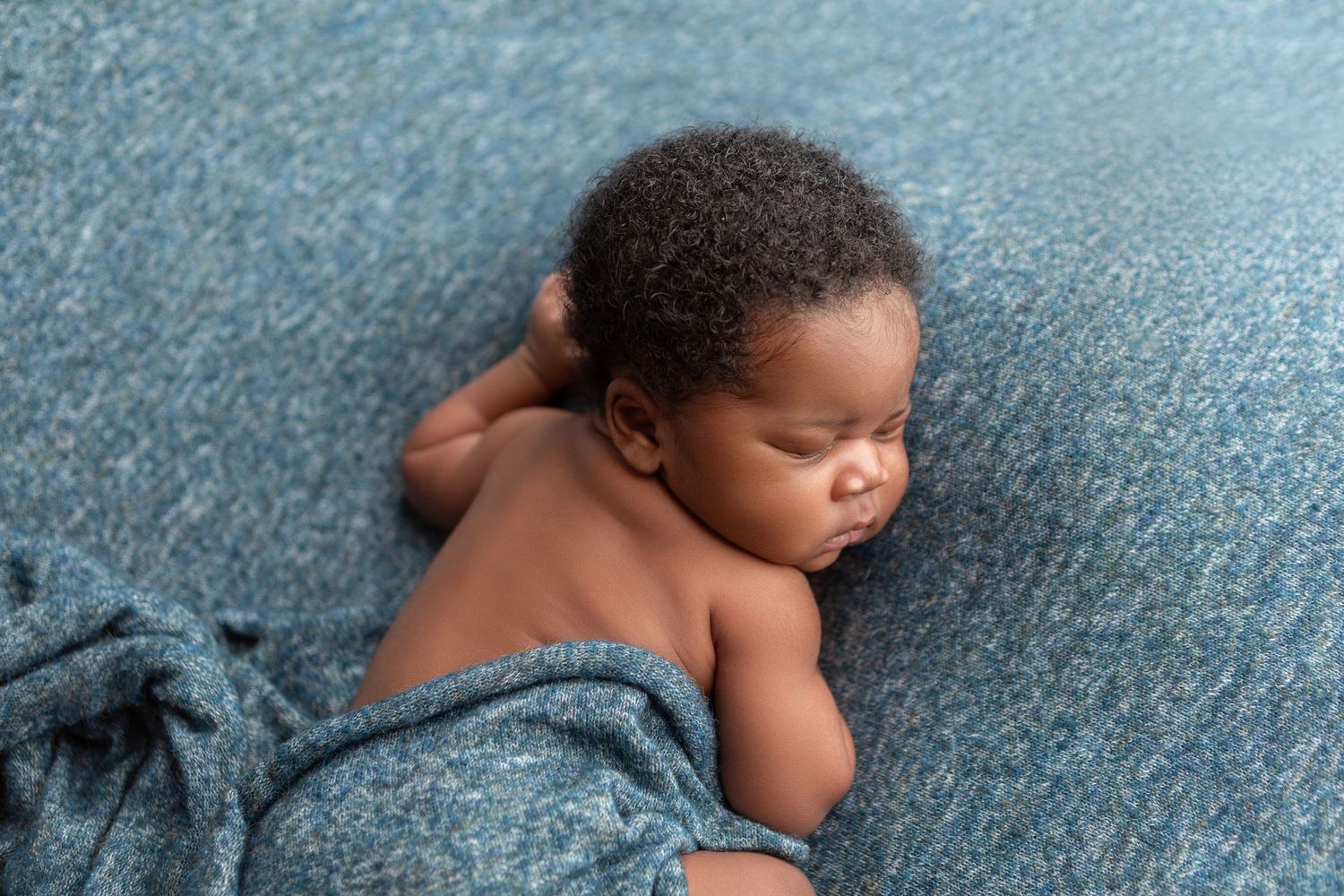 Newborn baby sleeping peacefully on a blue blanket, curled up in a natural pose during a professional newborn photography session.