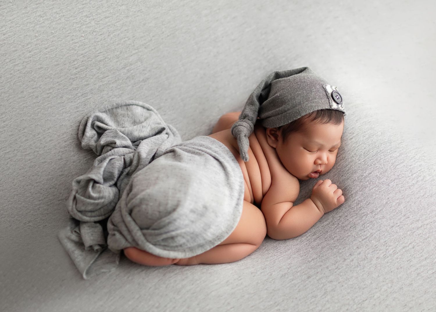 Sleeping newborn baby wrapped in a soft gray blanket wearing a knot hat on a white background.