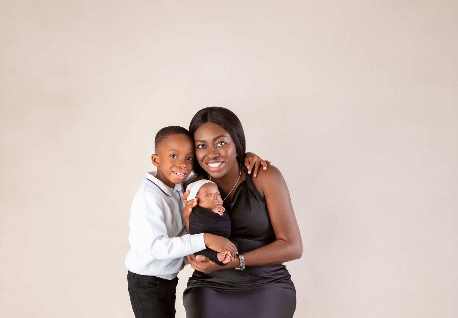 A smiling mother in a black dress holds her newborn while her young son hugs her in a studio portrait.