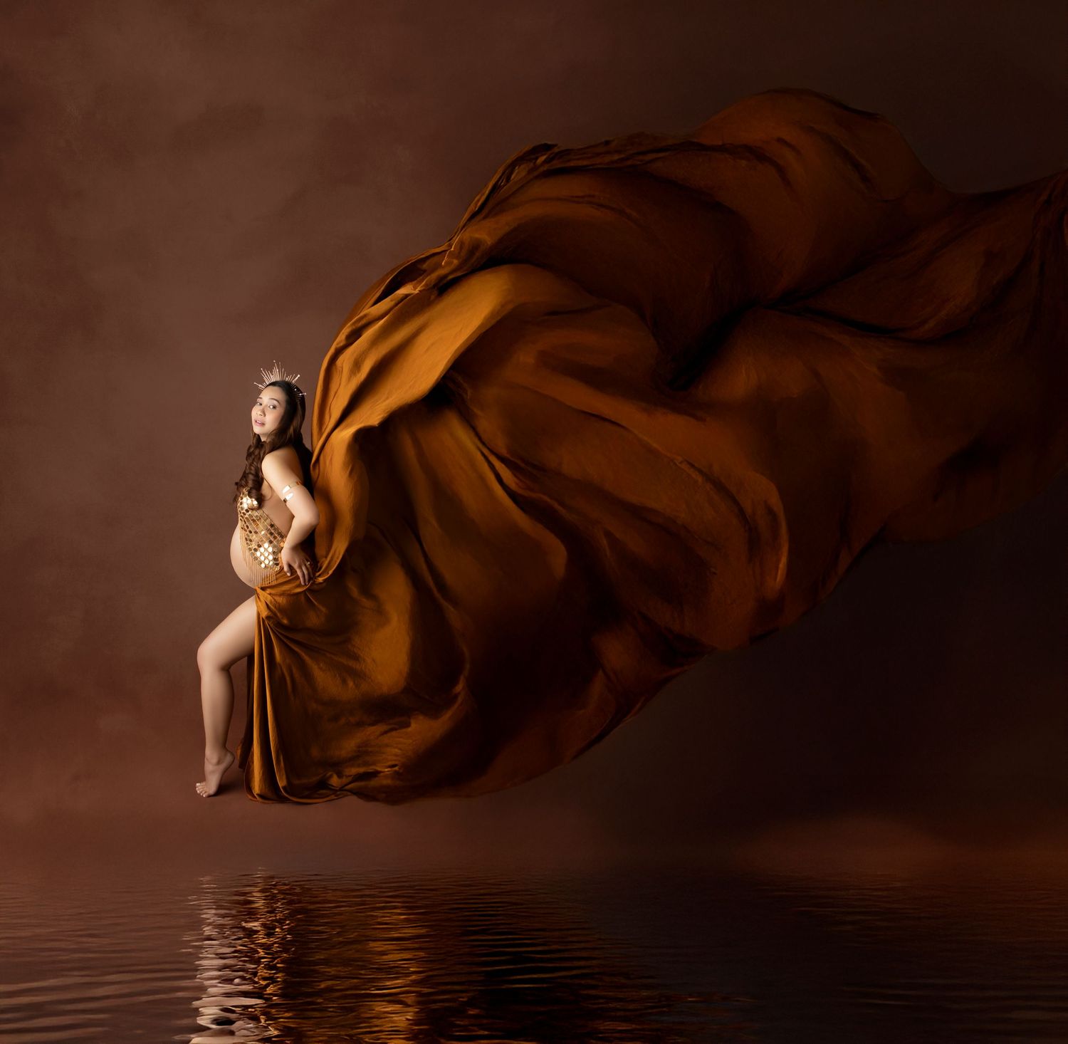 Pregnant woman in golden crown seated as amber silk fabric flows dramatically around her with water reflection.