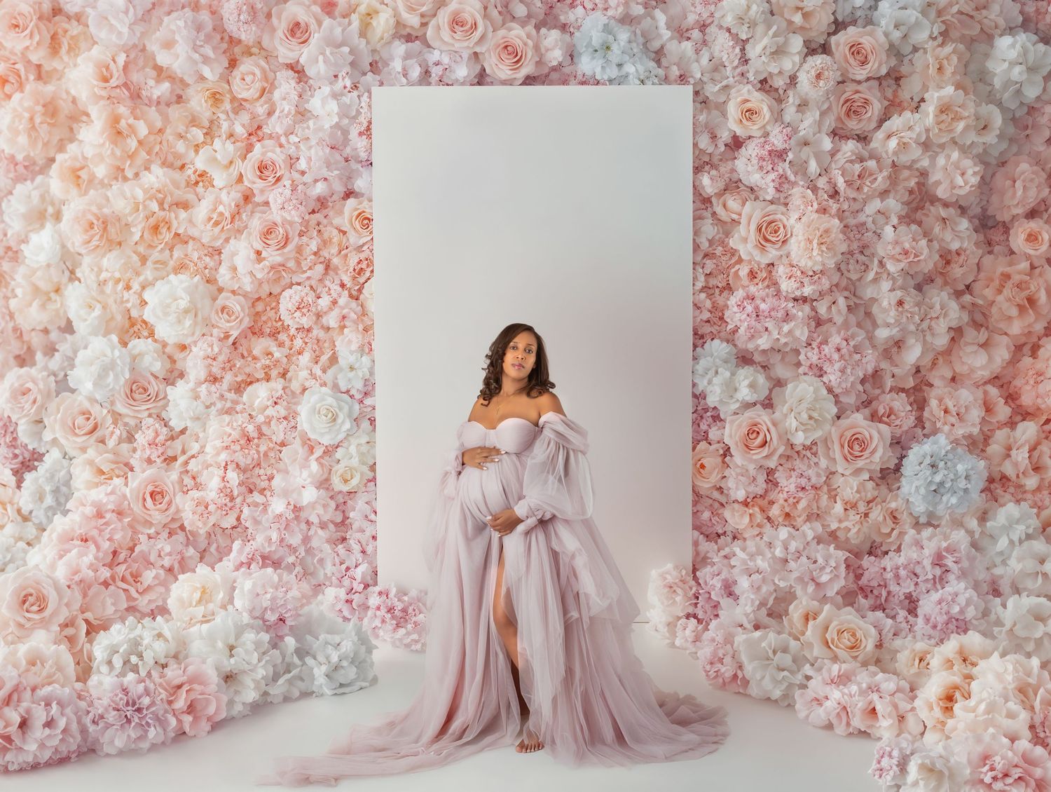 Maternity photo of woman in lavender off-shoulder gown standing before a wall of pastel pink and white flowers.