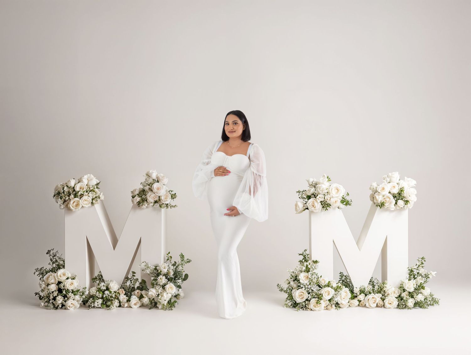 Pregnant woman in white jumpsuit poses between large floral M letters for a elegant maternity photo shoot.