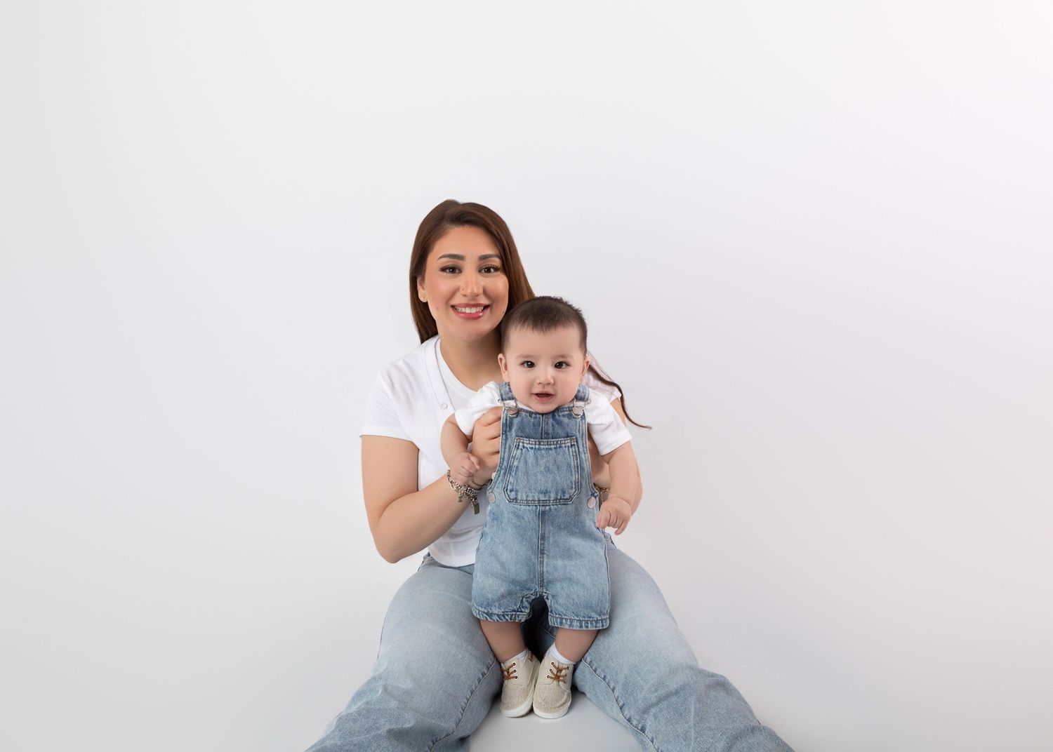 A smiling woman in white tee and jeans sits holding an adorable baby in denim overalls against white background.