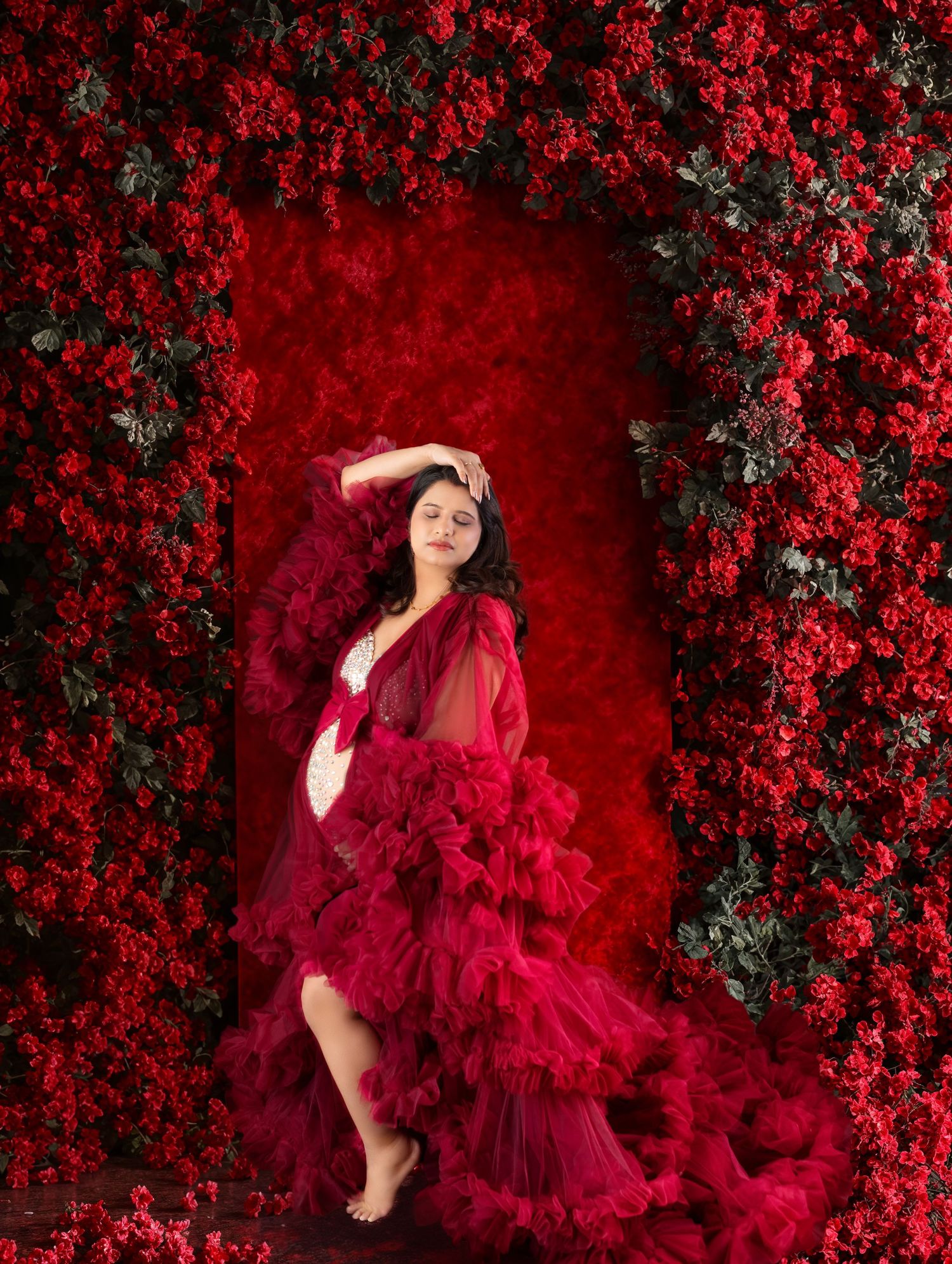 Pregnant woman in red tulle gown poses against lush red flower wall backdrop in elegant maternity portrait.