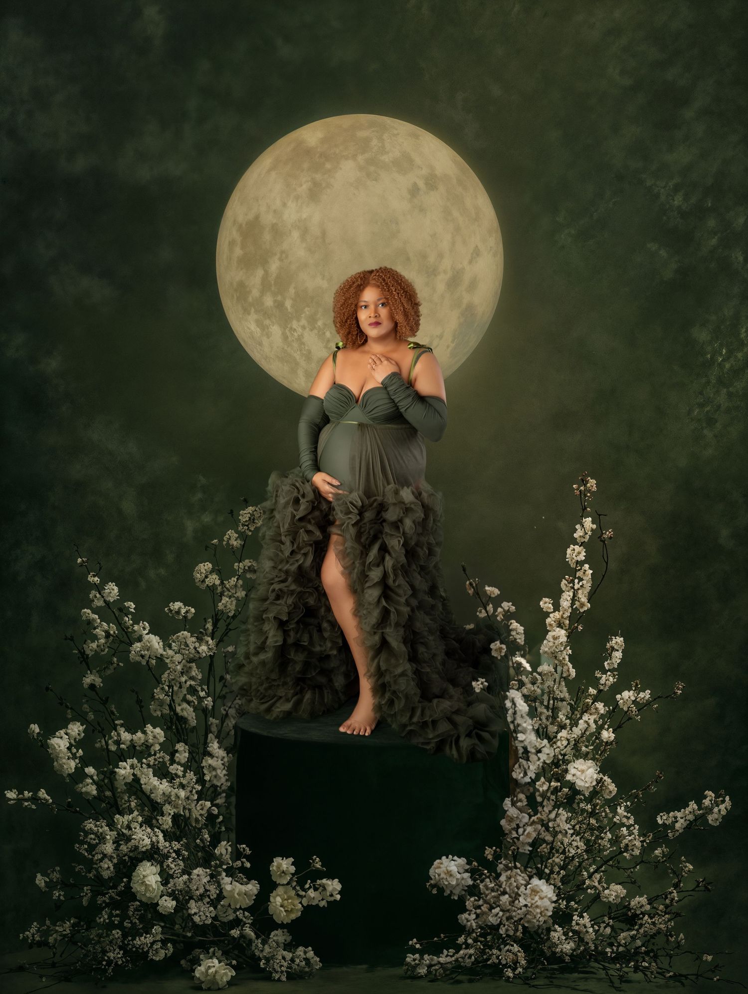 Pregnant woman in dark green ruffled gown stands on pedestal before glowing moon with white flowers.