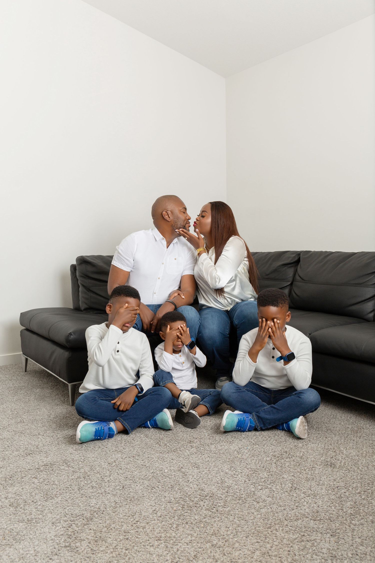 Family portrait with parents kissing on sofa while three young boys cover their eyes on the floor.