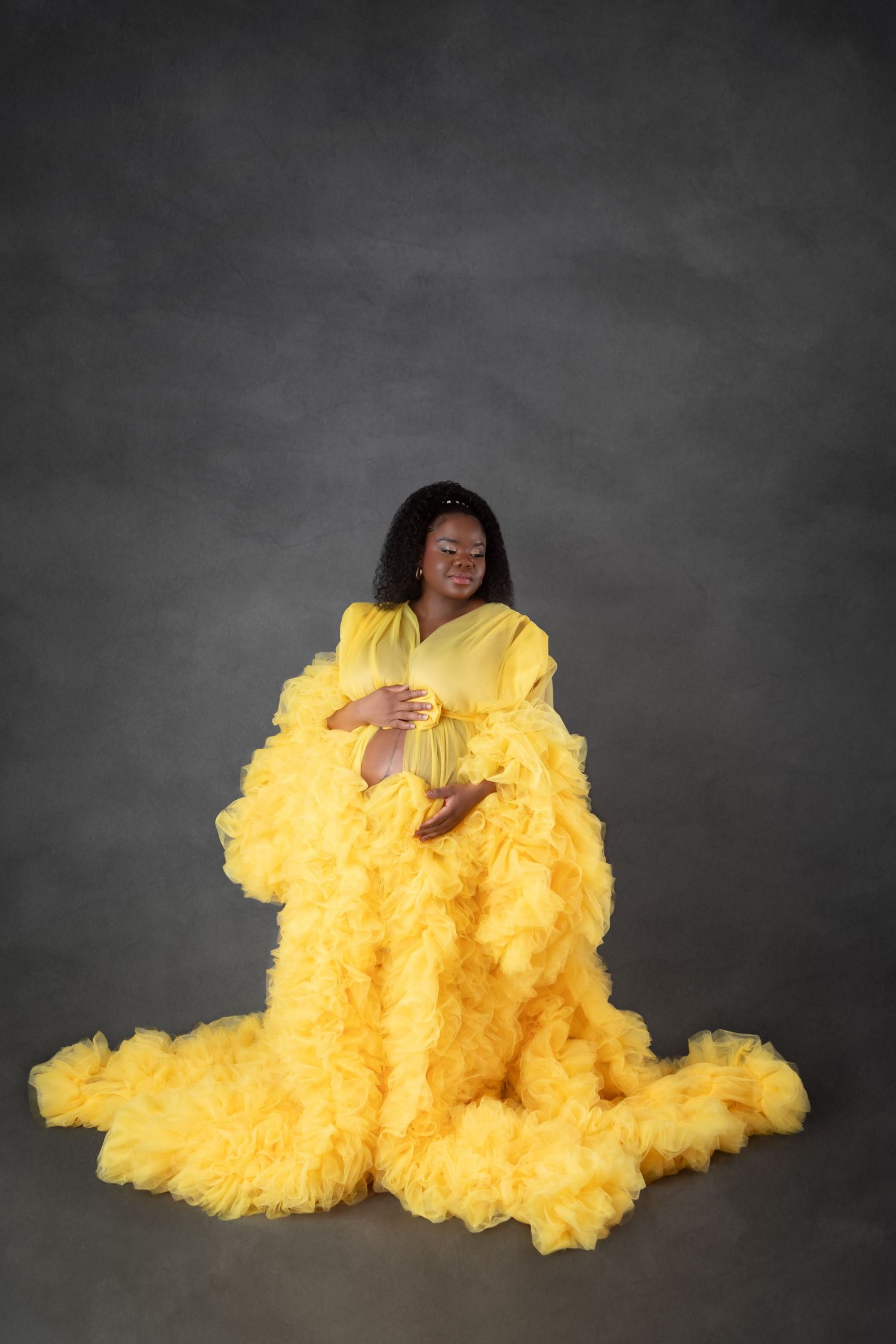 Pregnant woman in a voluminous yellow ruffled tulle maternity gown posing on a gray studio background.