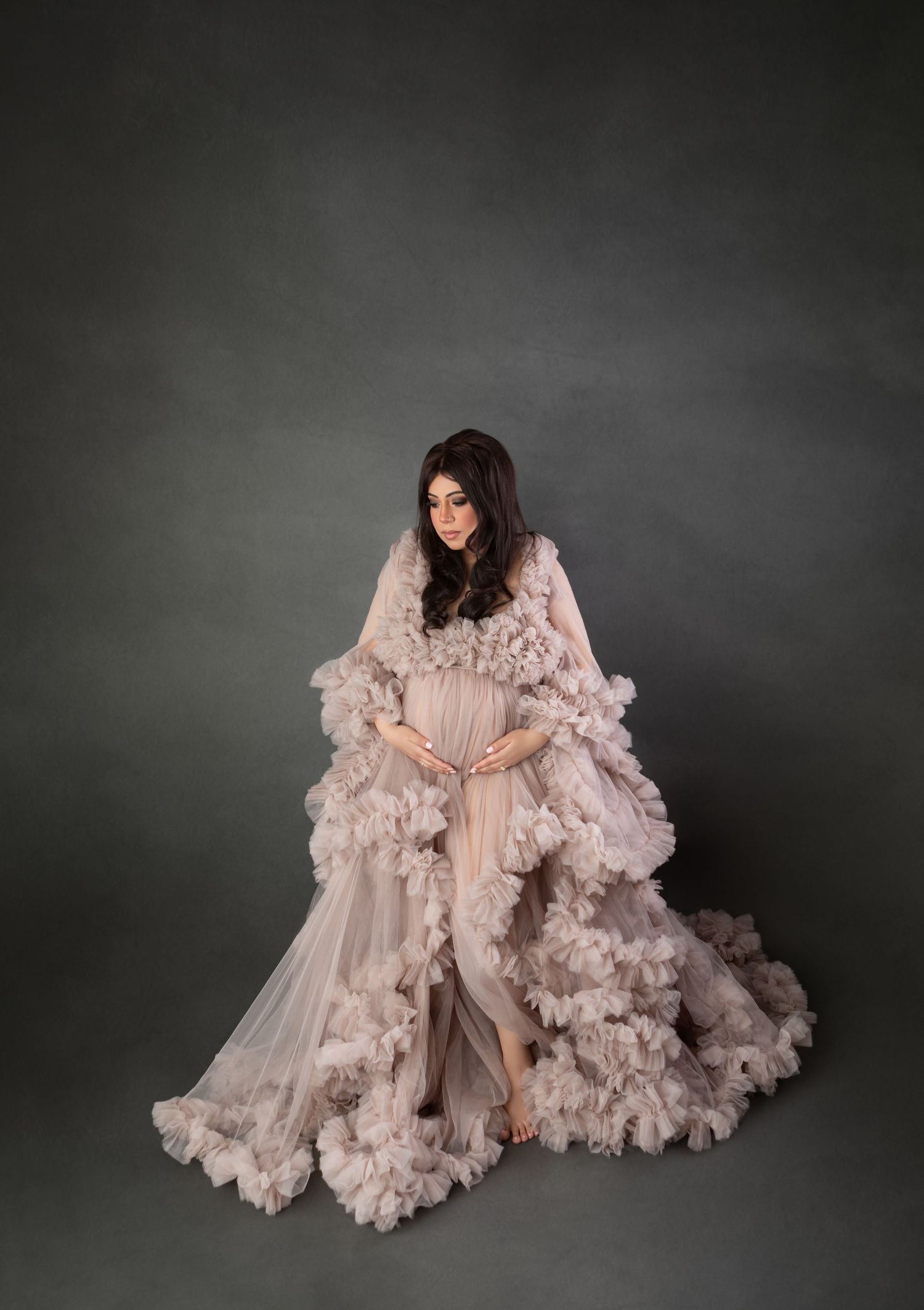 Pregnant woman in elegant blush ruffled tulle maternity gown posing in studio.