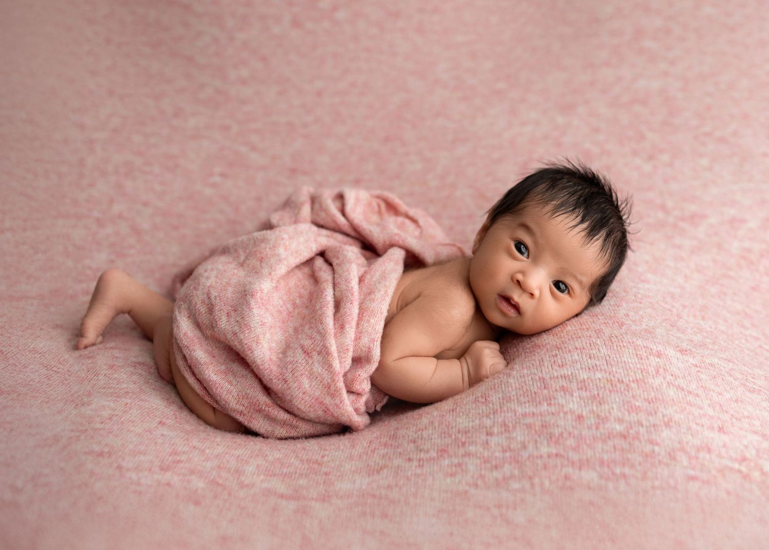 Awake newborn baby wrapped in a pink blanket lying on a soft pink textured backdrop.