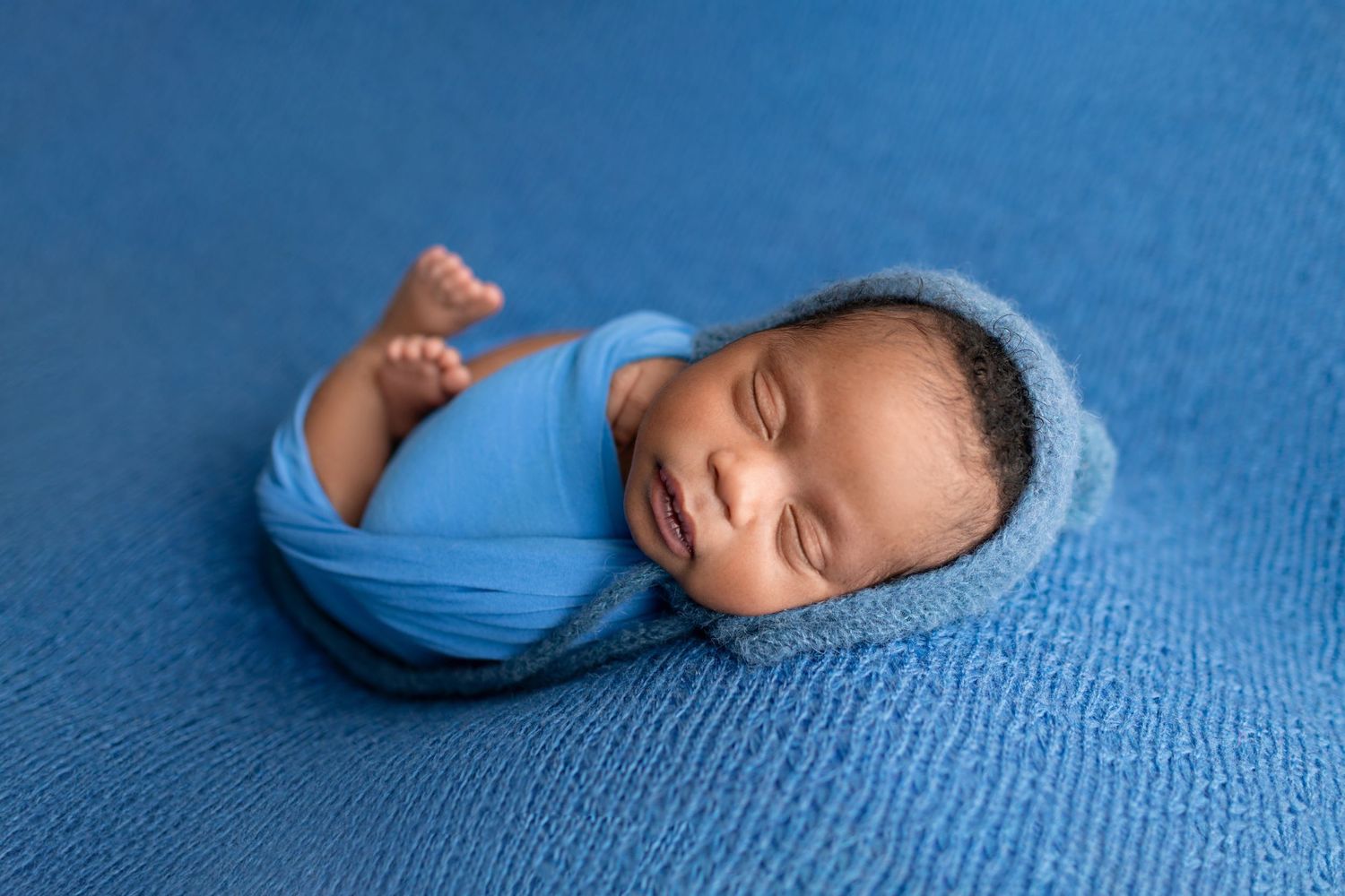 Smiling newborn baby swaddled in blue wrap lying on blue textured backdrop.