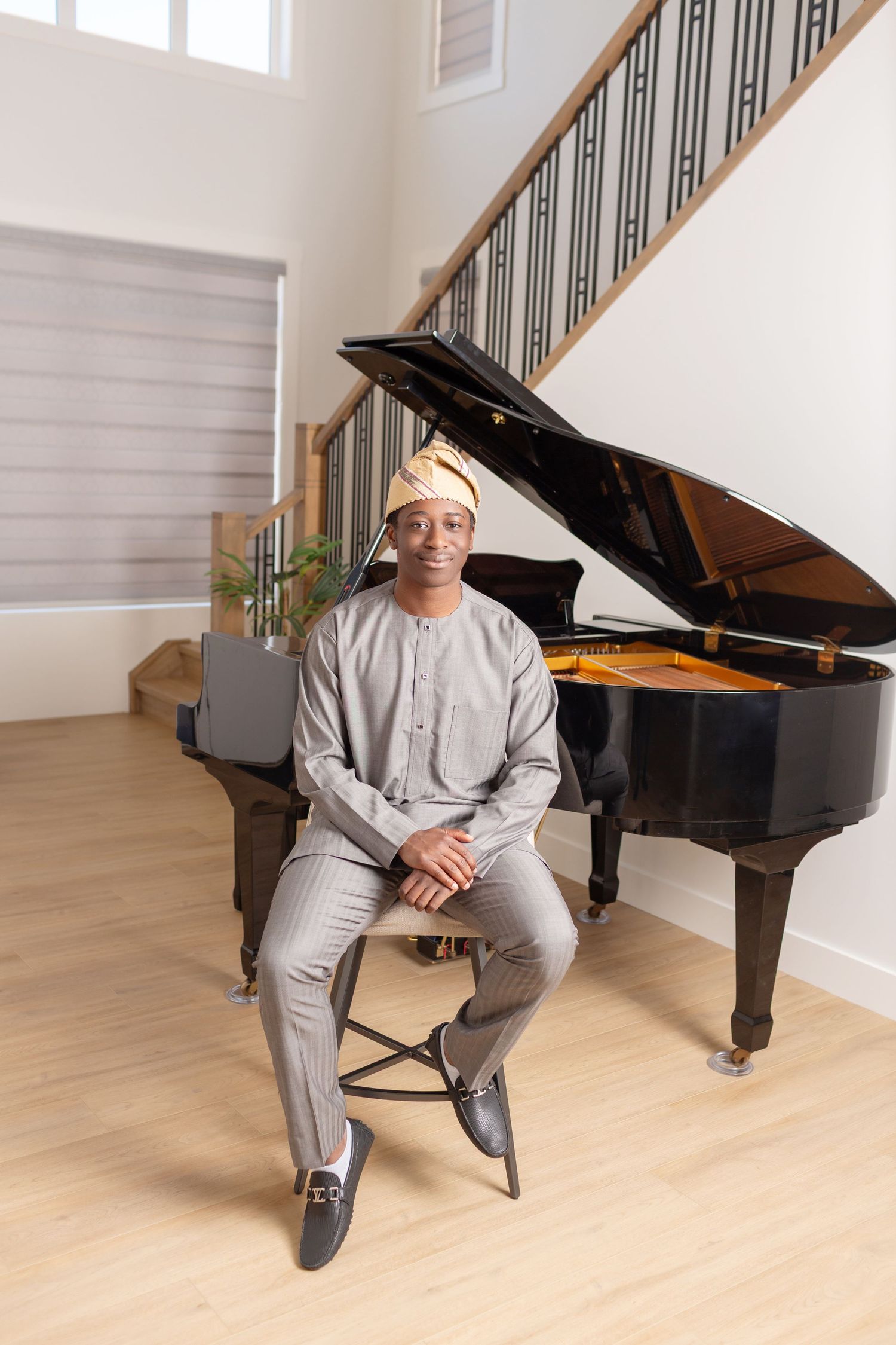 A man in traditional African attire and head wrap sits on a stool in front of a grand piano in an elegant home.
