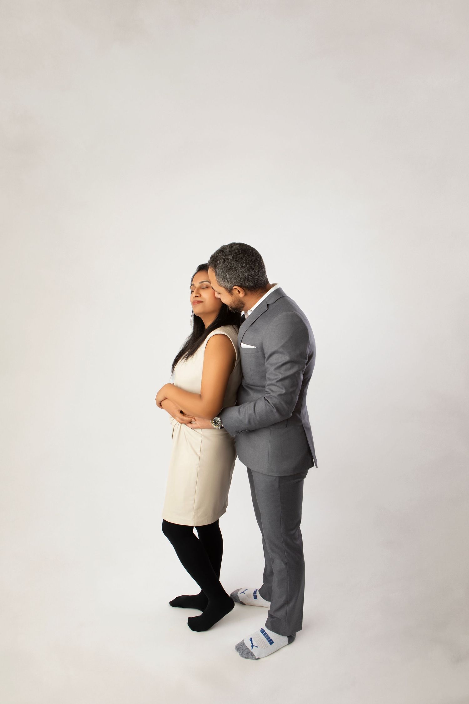 A couple in formal wear sharing a tender embrace, the man kissing the womans cheek against a white backdrop.