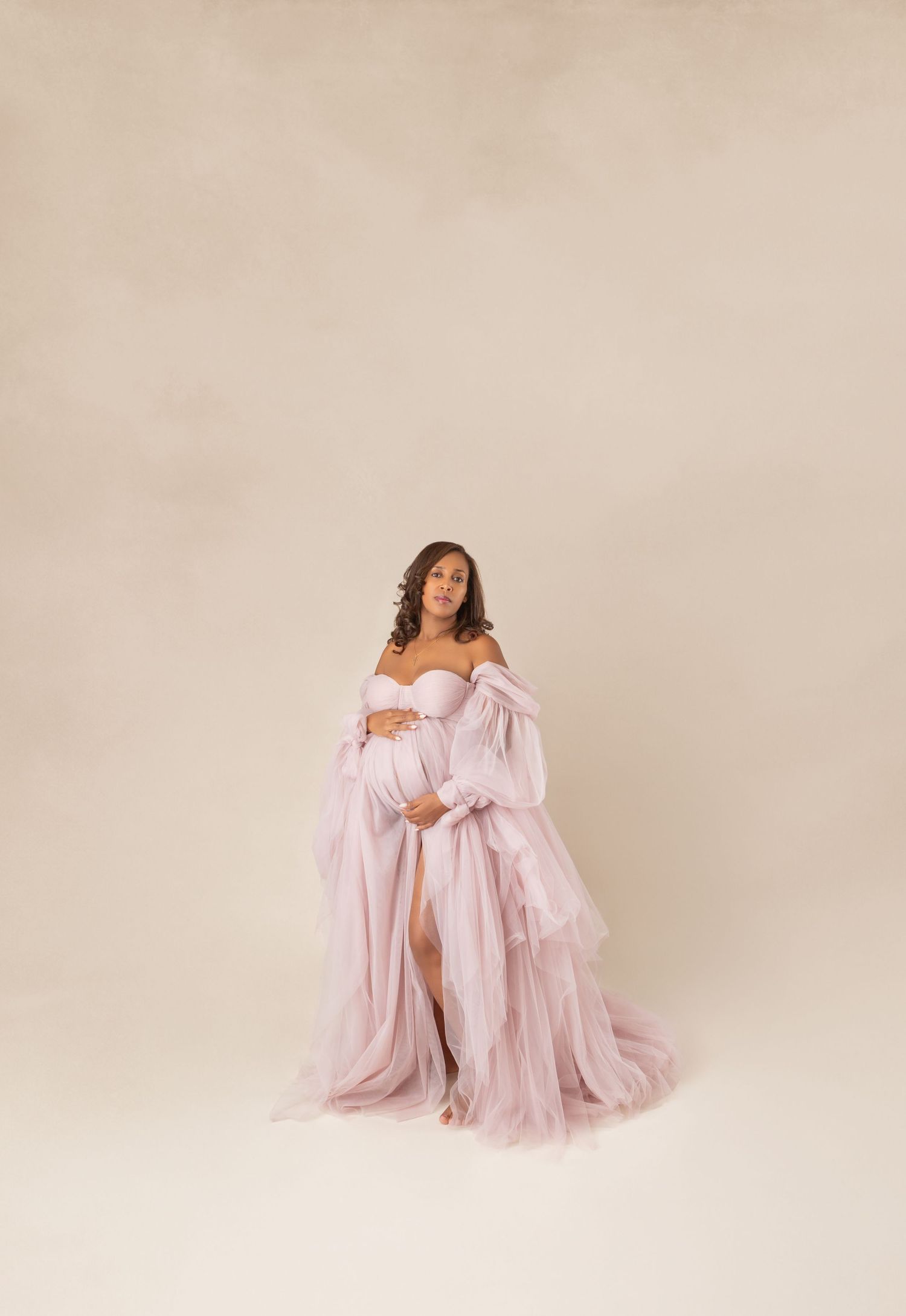 Pregnant woman in elegant pink tulle off-shoulder maternity gown posing in studio.