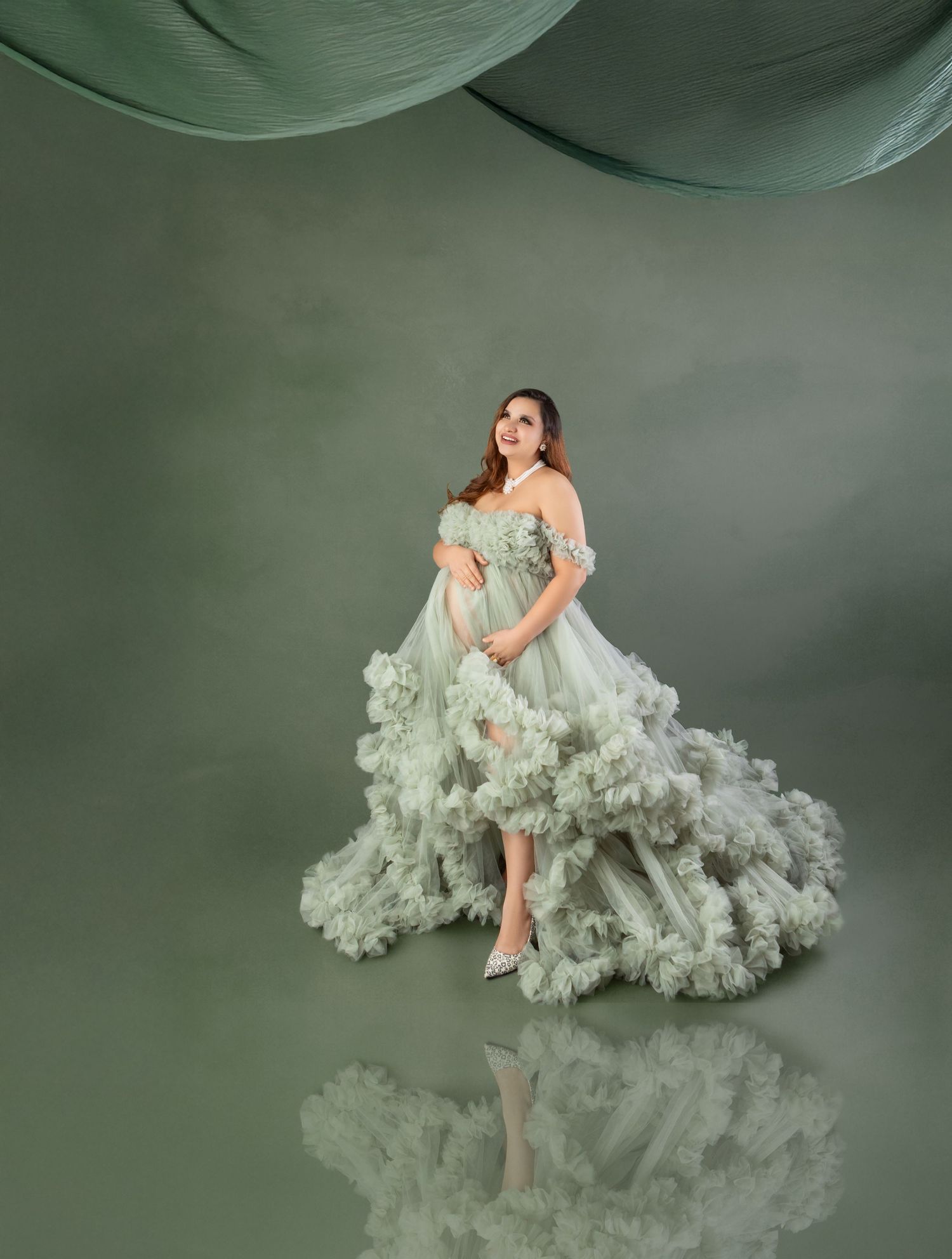Pregnant woman in sage green ruffled tulle maternity gown posing elegantly in a studio.