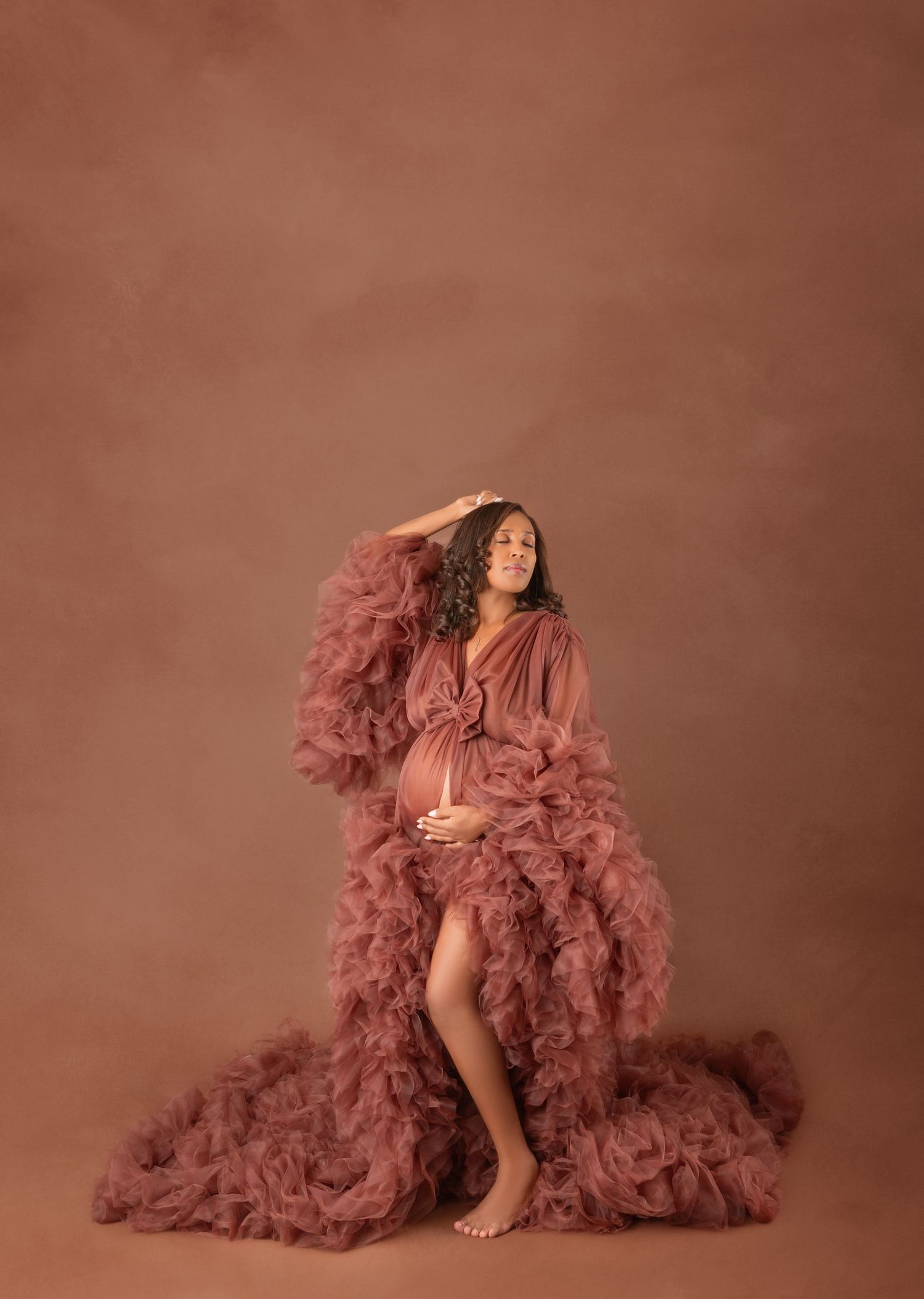 Pregnant woman in dusty rose ruffled tulle gown cradles bump against warm brown studio backdrop.