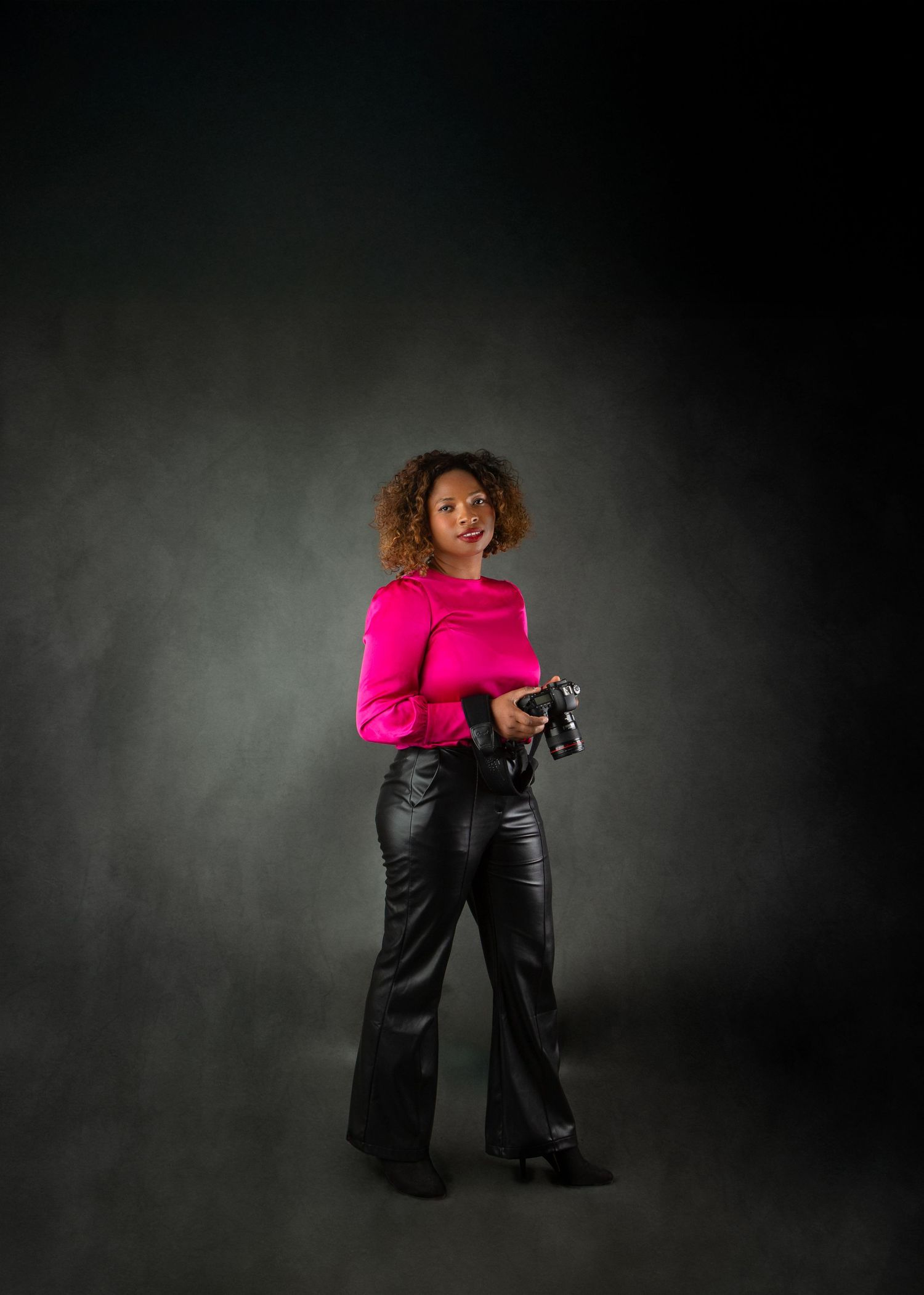 Woman in pink top and black leather pants holding camera in dark studio portrait.