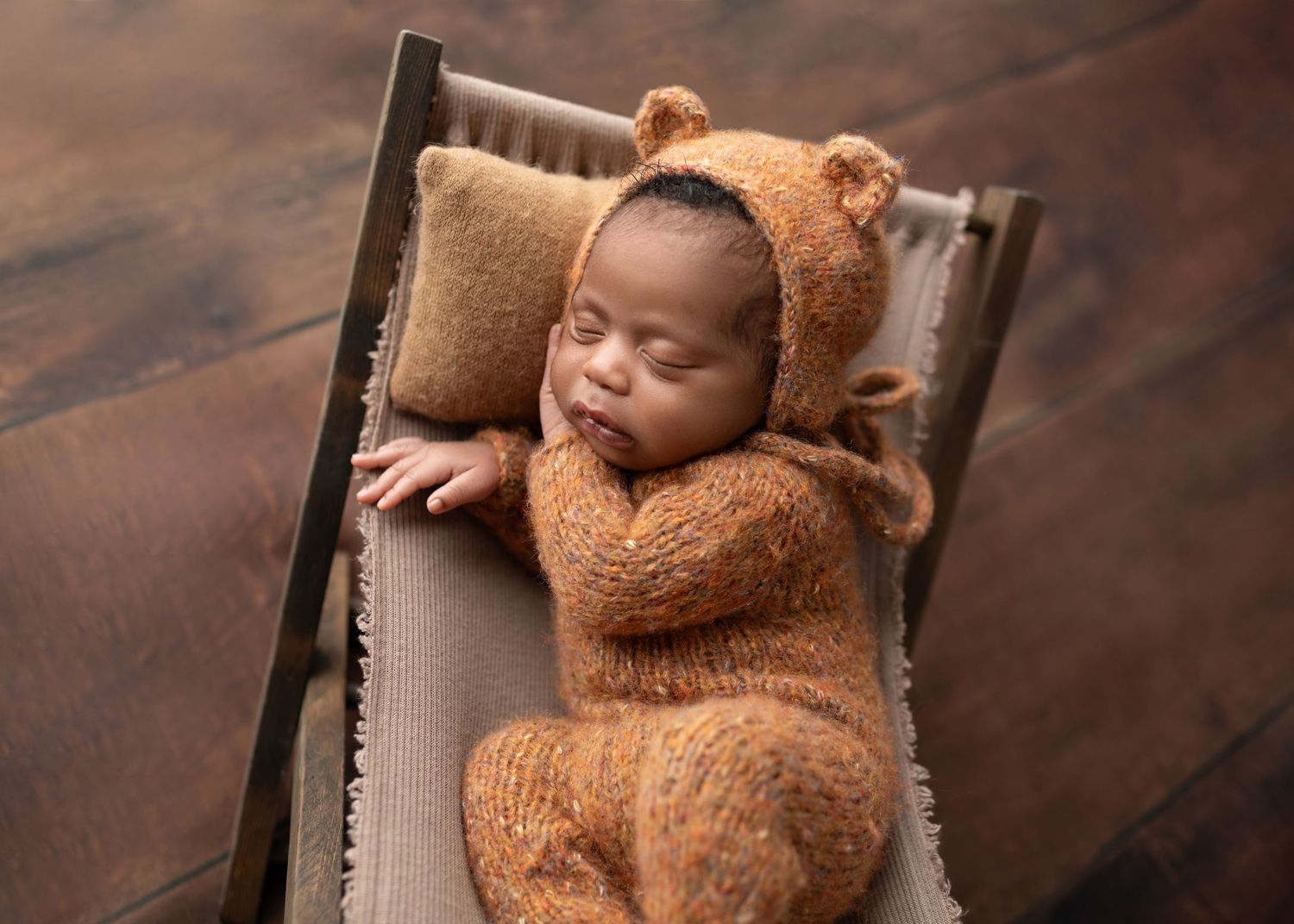 Adorable newborn in brown bear knit outfit napping on miniature wooden bed with pillow.